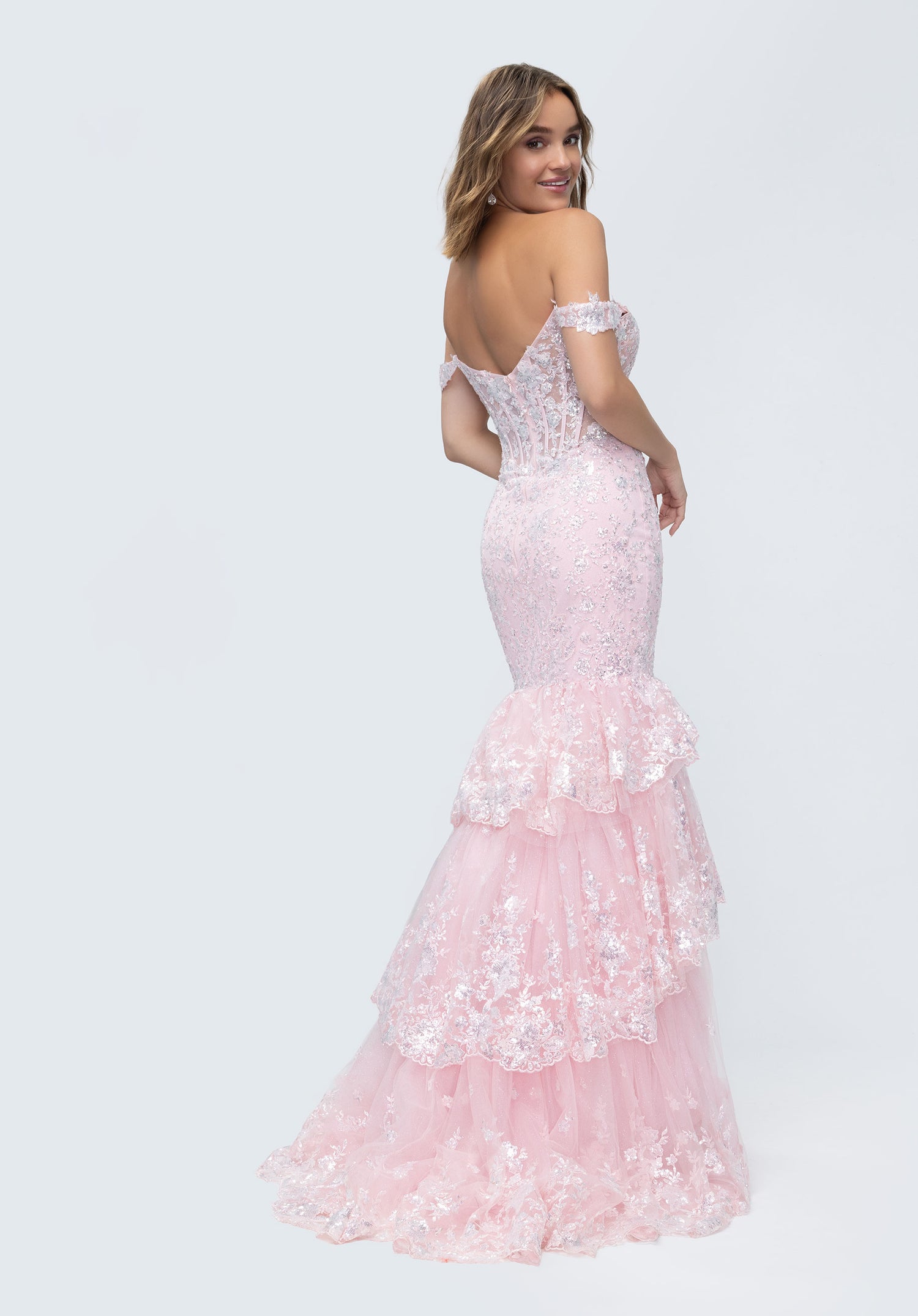 A woman in the Morilee 49274 long prom dress, a light pink off-shoulder lace corset mermaid gown with layered tulle ruffles, stands looking over her shoulder.