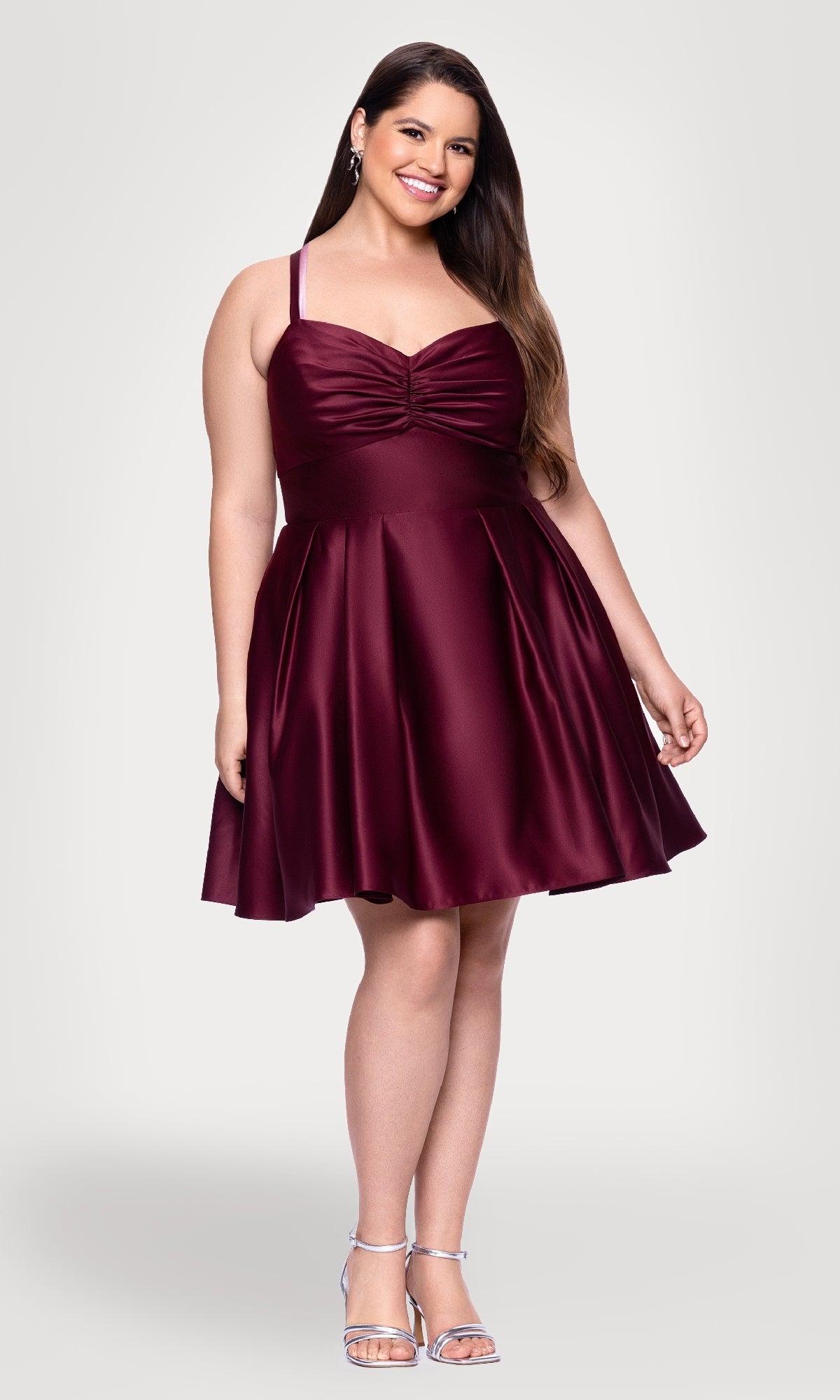 A woman smiles against a plain light background wearing the Plus Short Wine Hoco Dress Blondie Nites 4927BNW, featuring thin straps and burgundy satin, paired with silver heels.