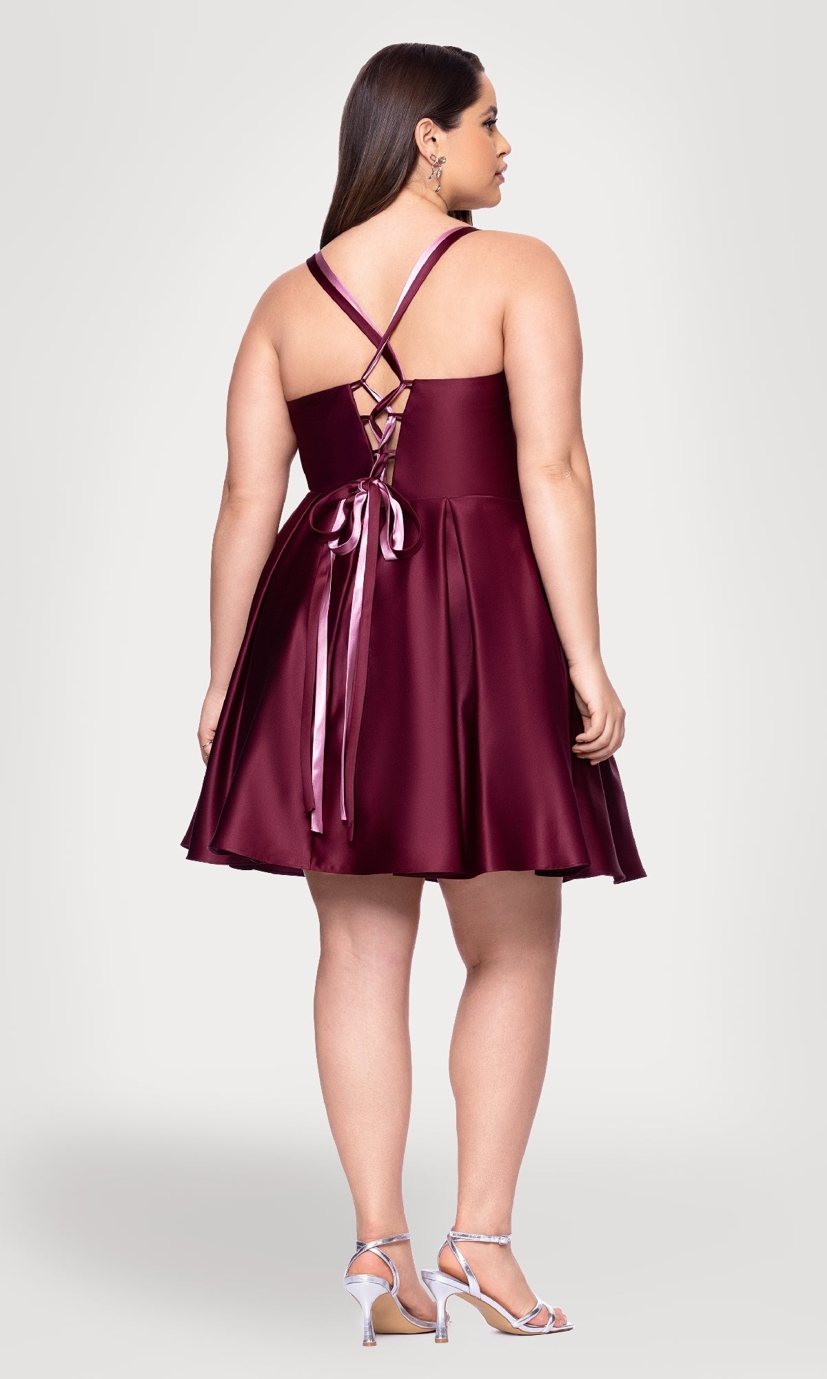 A woman wears the Plus Short Wine Hoco Dress Blondie Nites 4927BNW, featuring a crisscross lace-up back and satin ribbons, paired with silver heels; she faces away from the camera in this elegant party look.