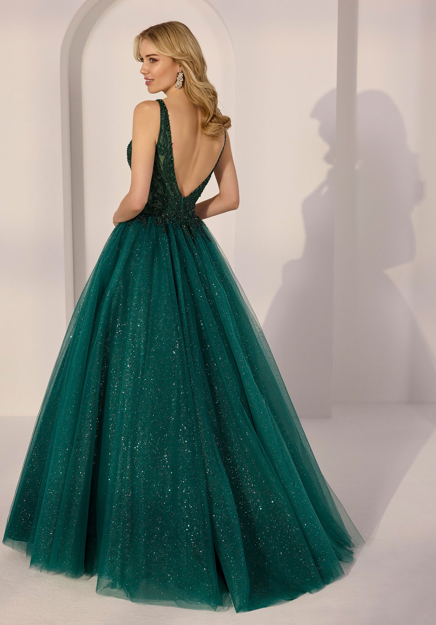 Woman wears the Morilee 49290 Long Prom Dress, a floor-length emerald gown with a deep V-back and a glittering tulle skirt, standing in a minimal white room with arched decor.