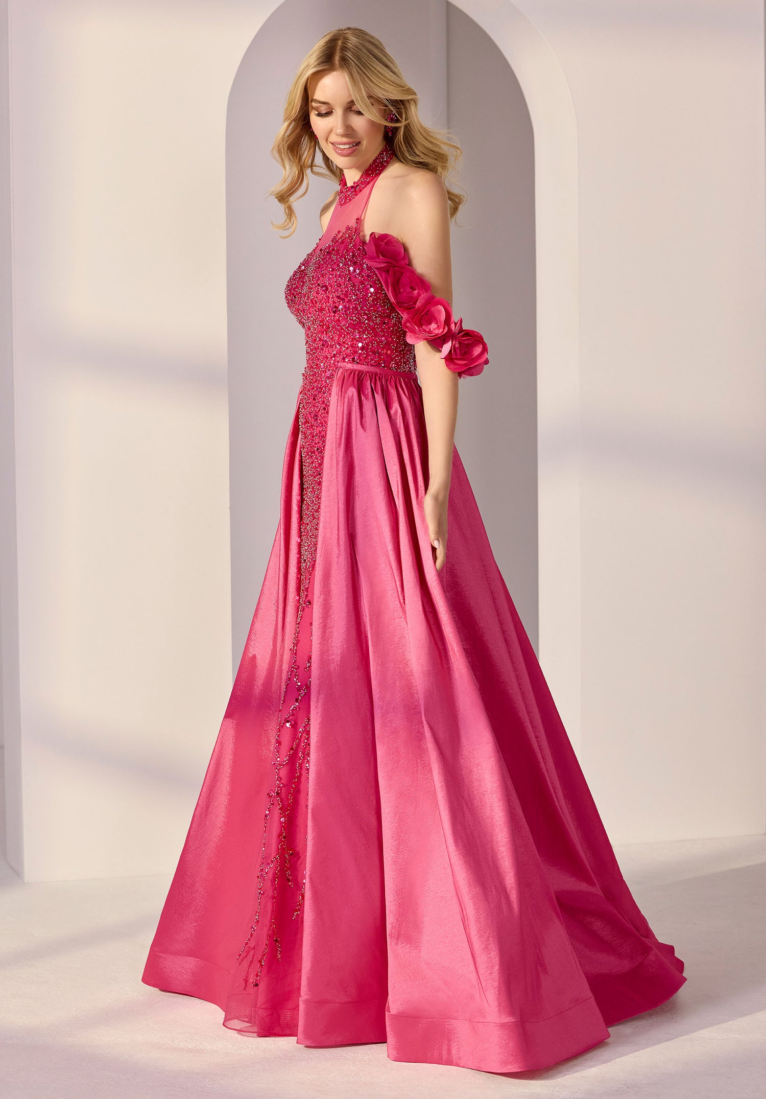 Indoors, a woman wears the Morilee 49313 long prom dress: a bright pink floor-length gown with floral shoulder details, sparkling bodice embellishments, and an elegant a-line overskirt.