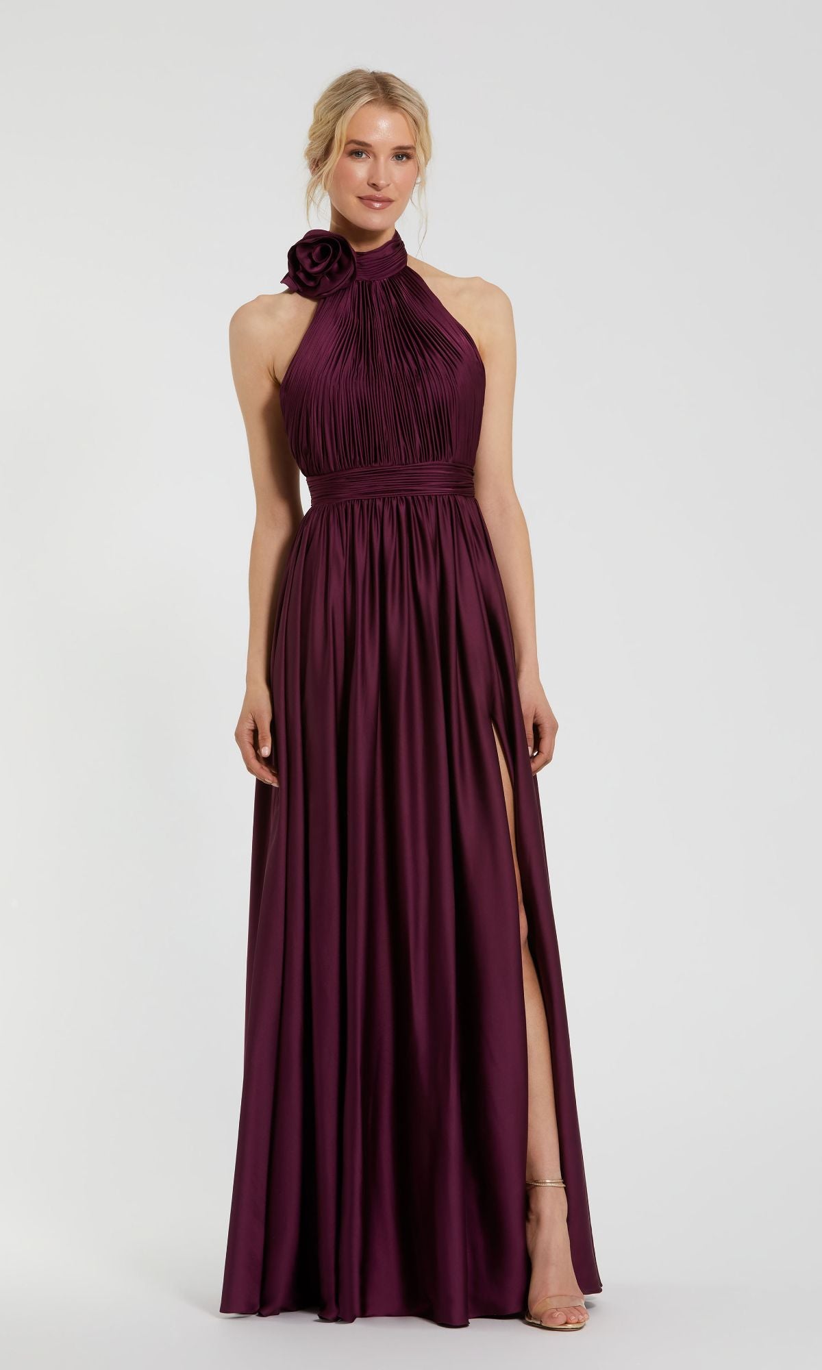 A woman wears the Mac Duggal 49333 high-neck halter formal dress—sleeveless, floor-length, in dark purple—with a pleated bodice, floral shoulder detail, and a high side slit.