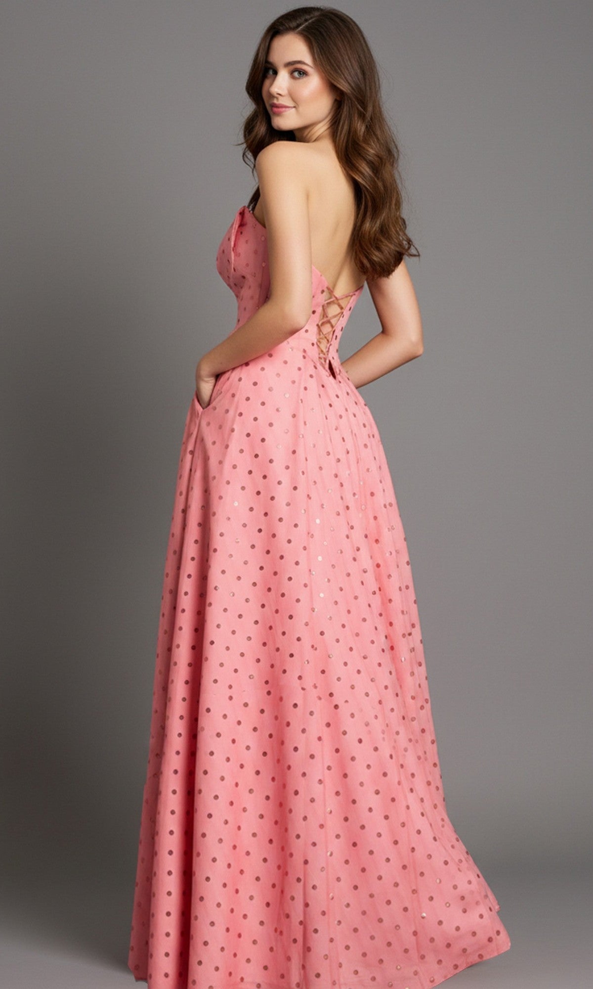 Woman wearing Jovani 49340, a pink floor-length prom dress with metallic polka dots, poses sideways against a plain gray background.