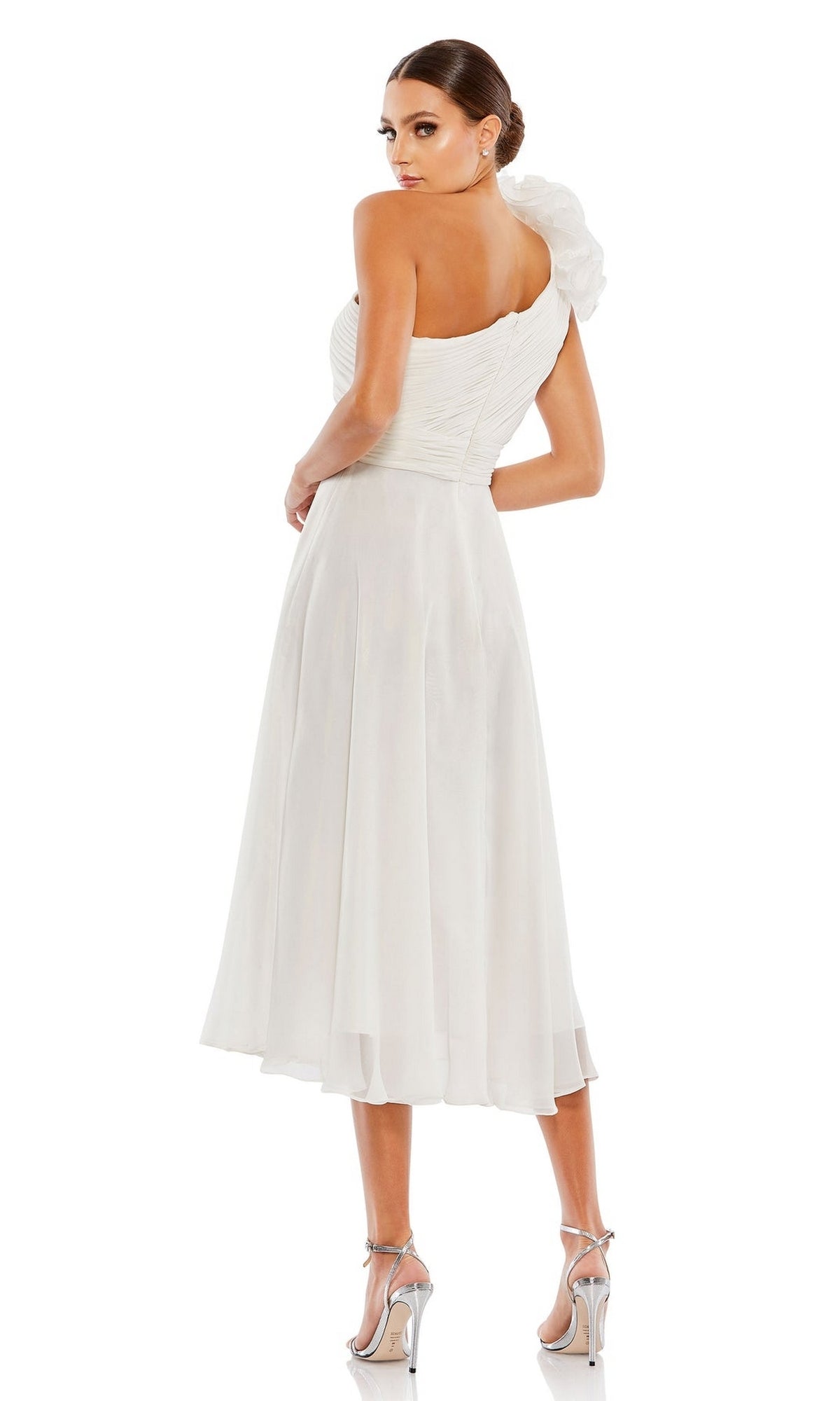 A woman stands facing away from the camera in the Mac Duggal 49491 knee-length white party dress, featuring a one-shoulder design with floral detail, paired with high-heeled sandals.