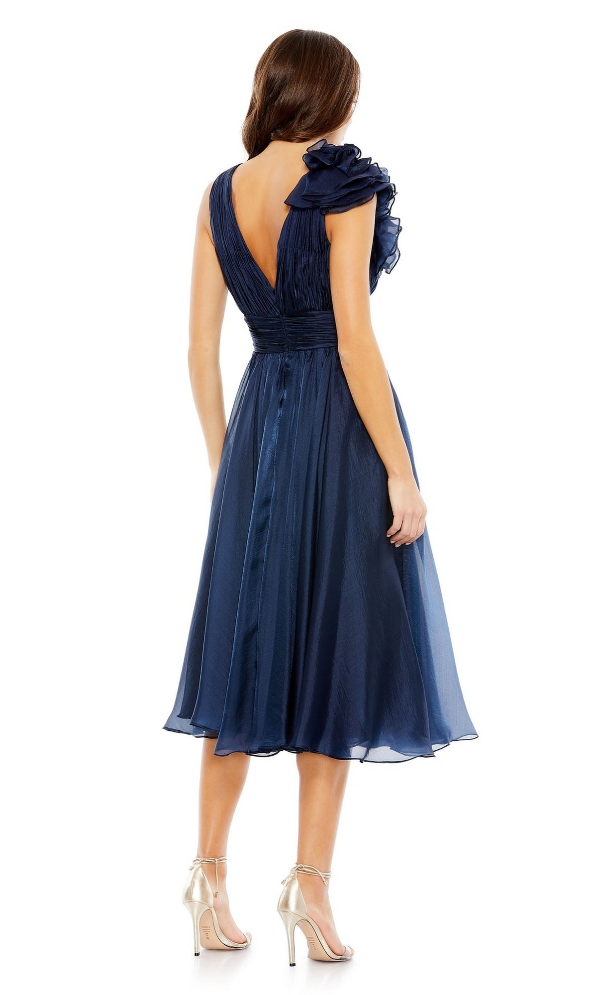 A woman in the Short Ruched Wedding-Guest Dress: Mac Duggal 49494, featuring navy blue fabric, ruffled shoulders, a V-back, and silver high-heeled sandals, stands facing away.