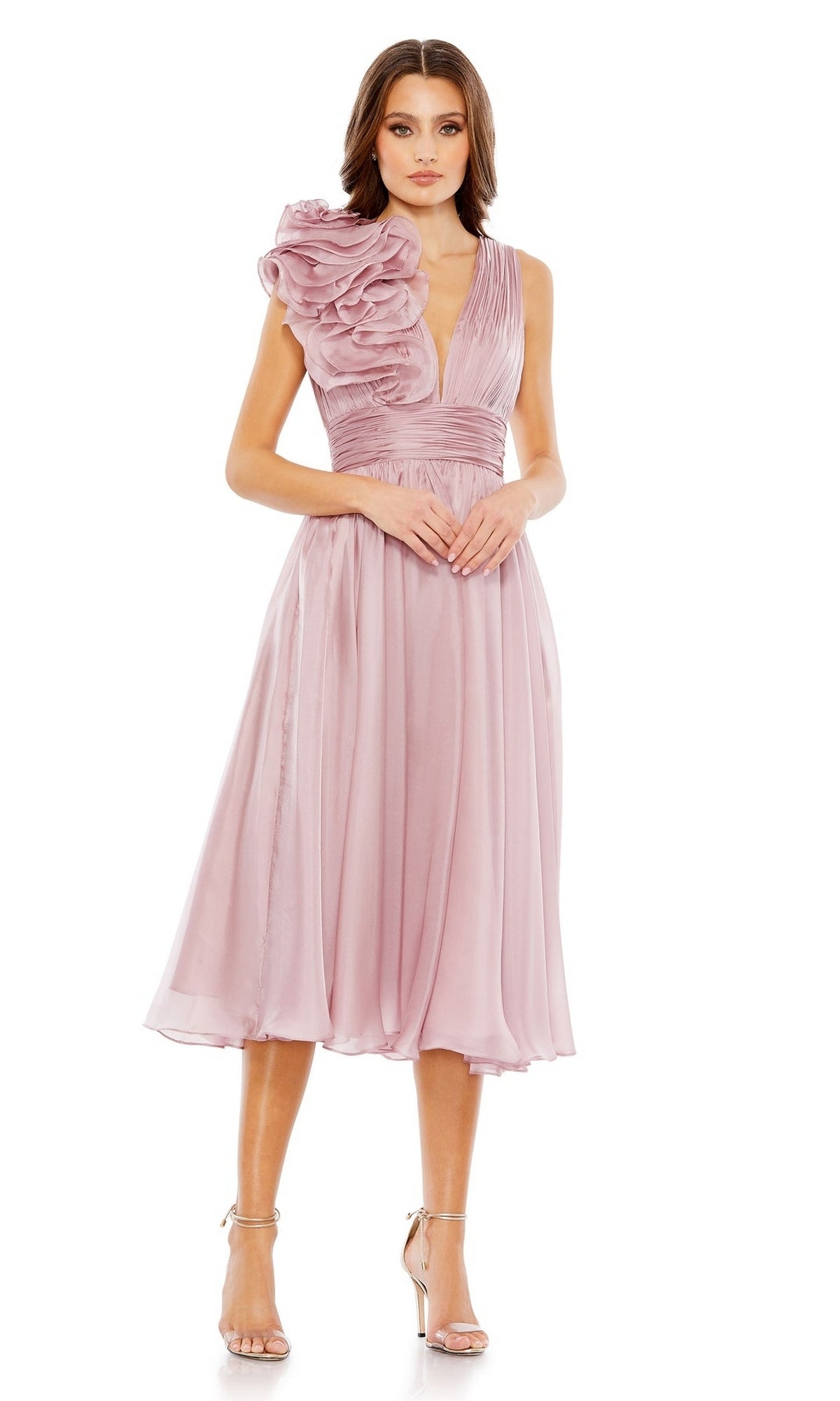 A woman stands wearing the Mac Duggal 49494 short ruched wedding guest dress, a sleeveless, knee-length dusty pink style with a large one-shoulder ruffle detail, paired with strappy high-heeled sandals.