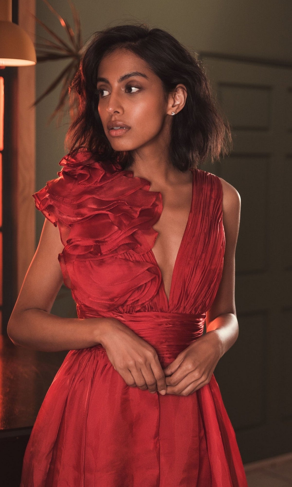 Wearing the Mac Duggal 49494, a short ruched wedding-guest dress with a deep neckline and one-shoulder ruffle, a woman stands indoors looking to the side—a stunning choice for any wedding celebration.