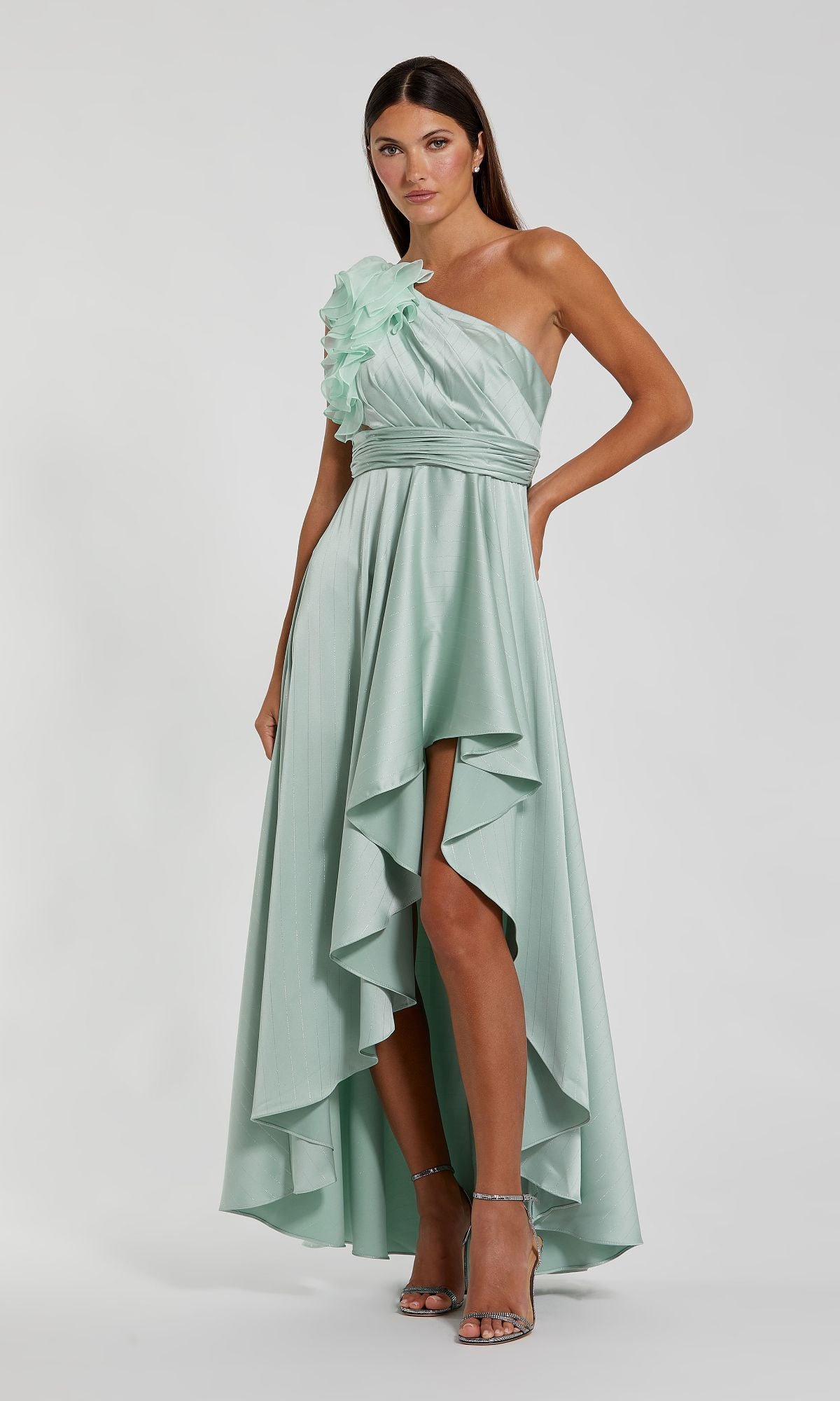 A woman models the Mac Duggal 49523 one-shoulder wedding guest dress in light green, featuring a high-low hem and floral ruffle detail, paired with strappy heels—a perfect party outfit.