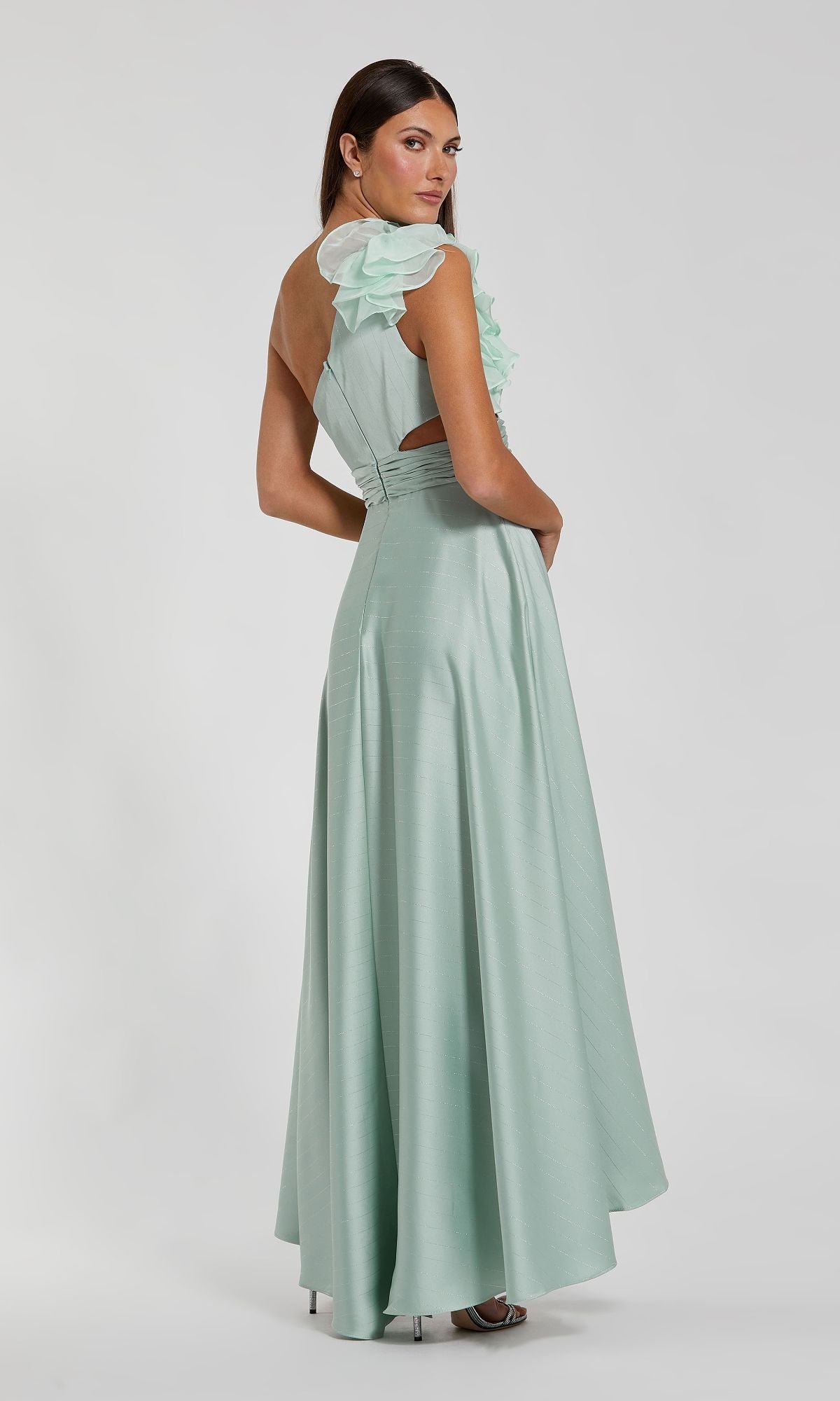 A woman faces away, wearing the Mac Duggal 49523 one-shoulder wedding guest dress with ruffle detail and a waist cutout, in light green, against a plain background.