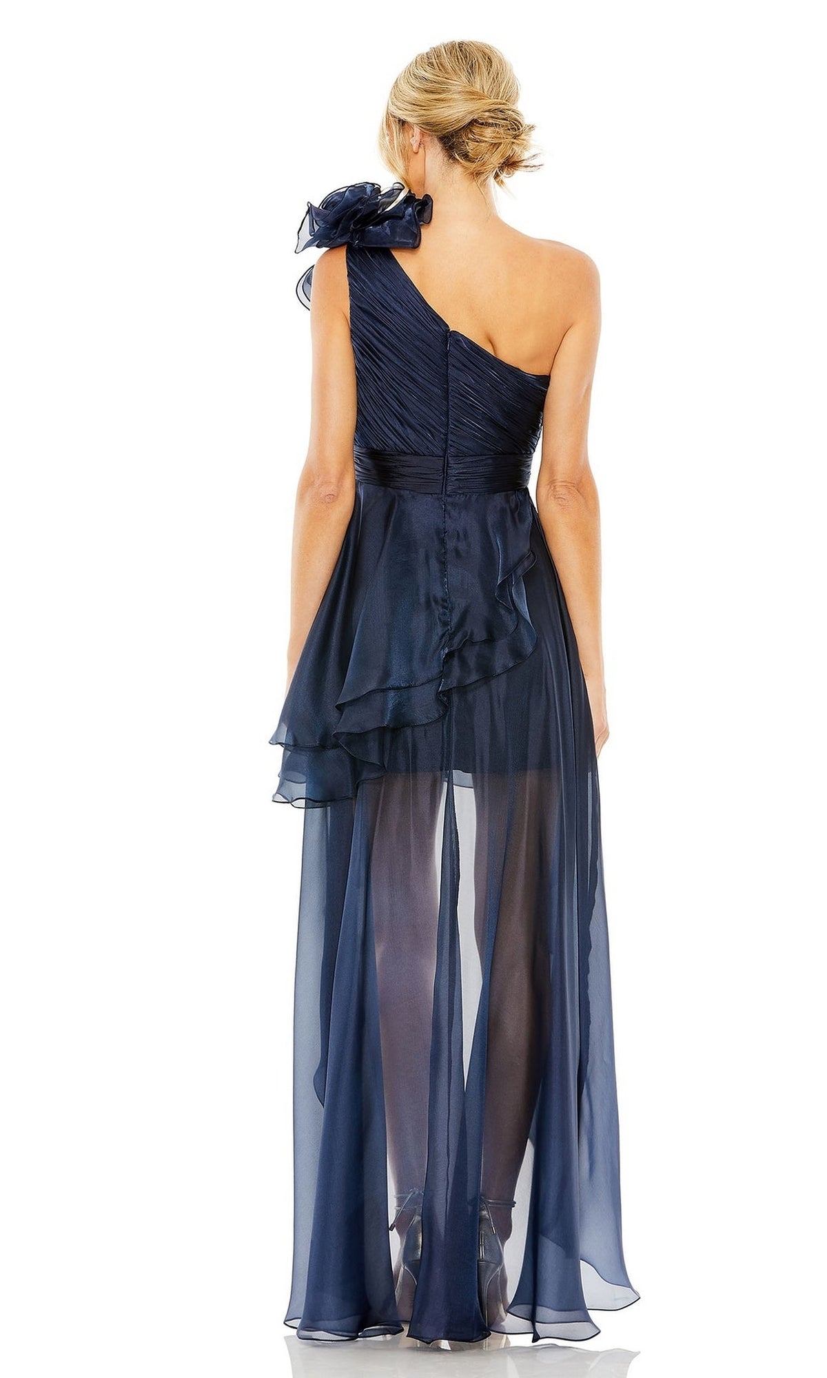 A woman, hair in a low bun, stands facing away in the Short Wedding Guest Dress 49527 by Mac Duggal—a midnight blue, one-shoulder party dress with a sheer layered gown, asymmetric hem, and textured bodice.