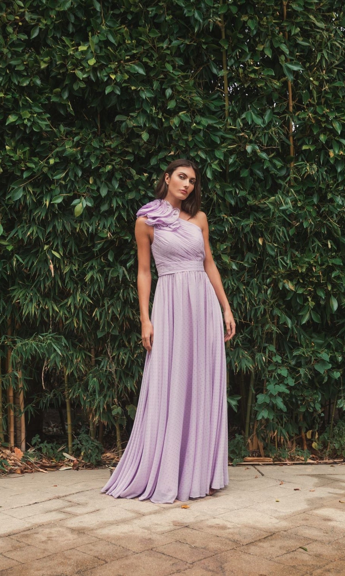 Woman in a long, light purple One-Shoulder Long Formal Dress: Mac Duggal 49536 stands on a paved path before dense green foliage.