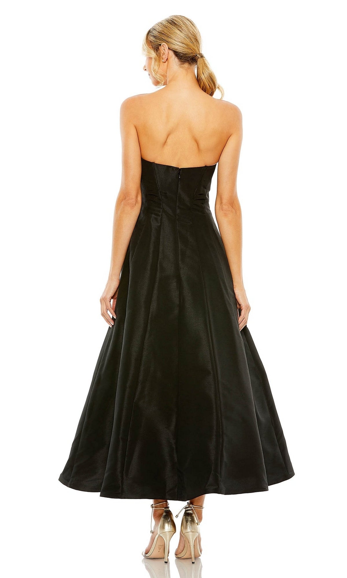 A blonde woman in a low ponytail wears the strapless black Mac Duggal 49605 tea-length wedding-guest dress and metallic heels, standing with her back to the camera against a white background.