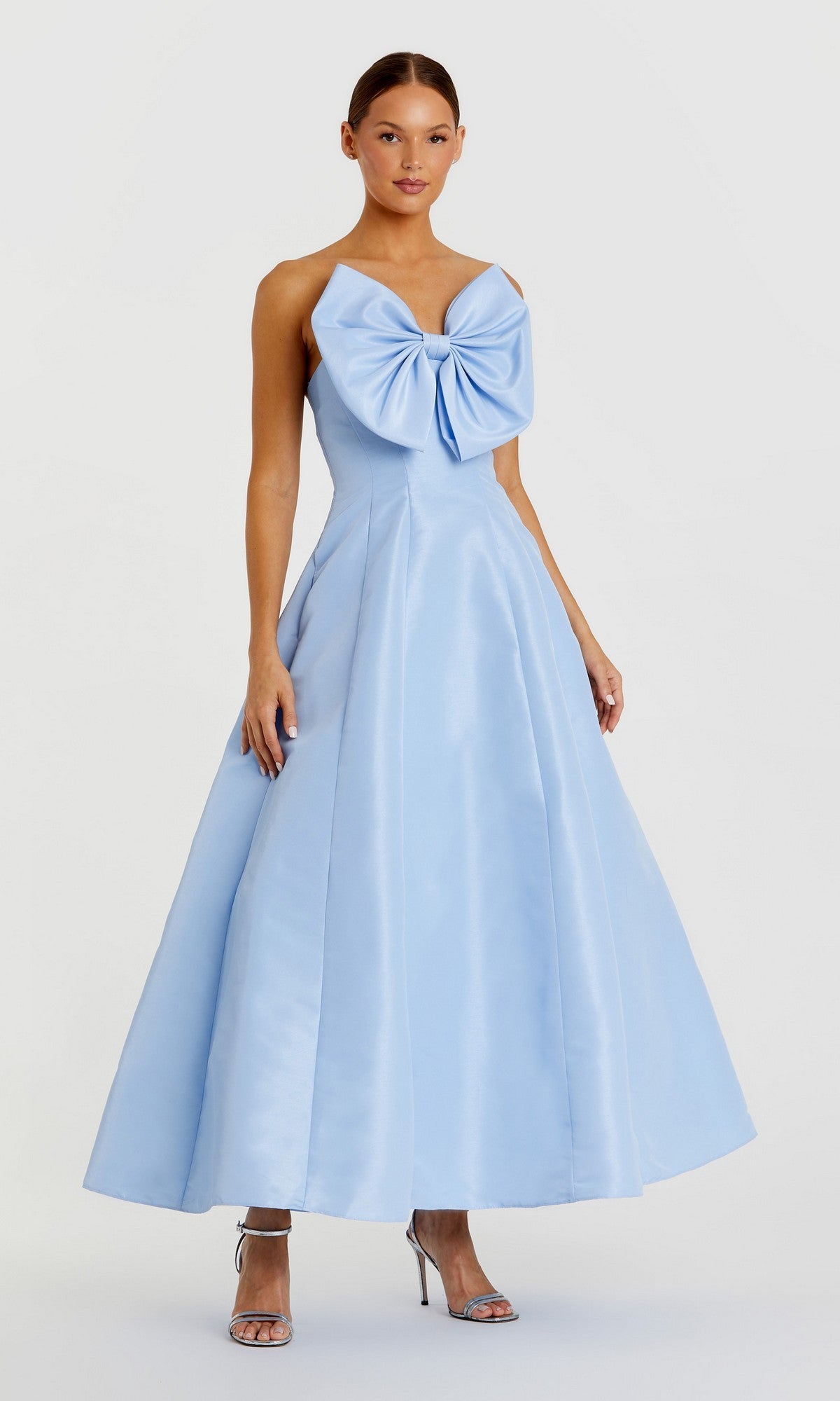 A woman in the Mac Duggal 49605 tea-length wedding-guest dress, light blue with a large chest bow, pairs silver high-heeled sandals while standing against a plain light background.