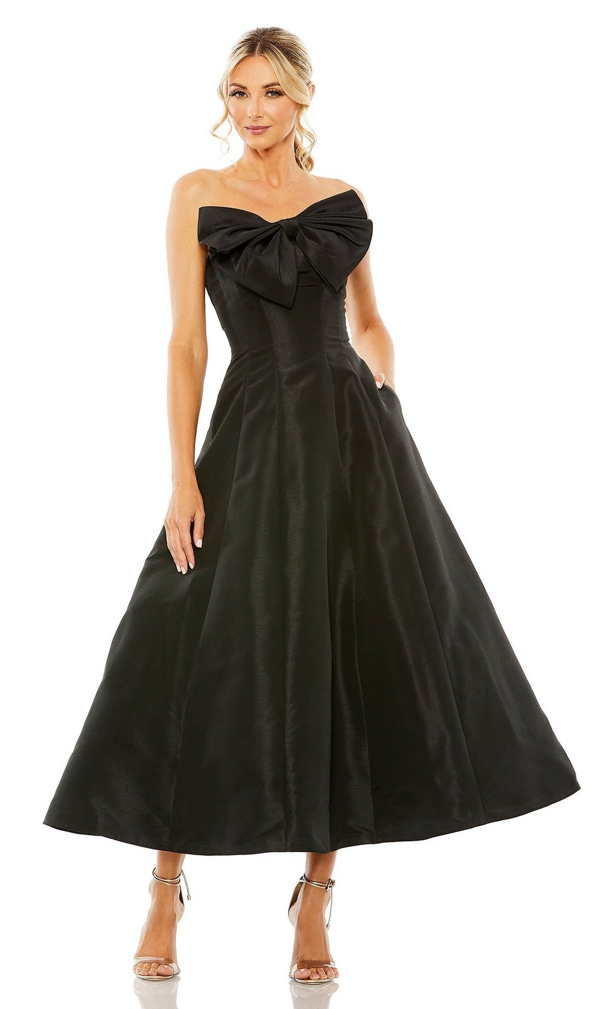 A woman wears the Mac Duggal 49605 tea-length wedding-guest dress—a strapless black midi with a large bow on the bodice—standing with one hand in her pocket and silver high-heeled sandals.