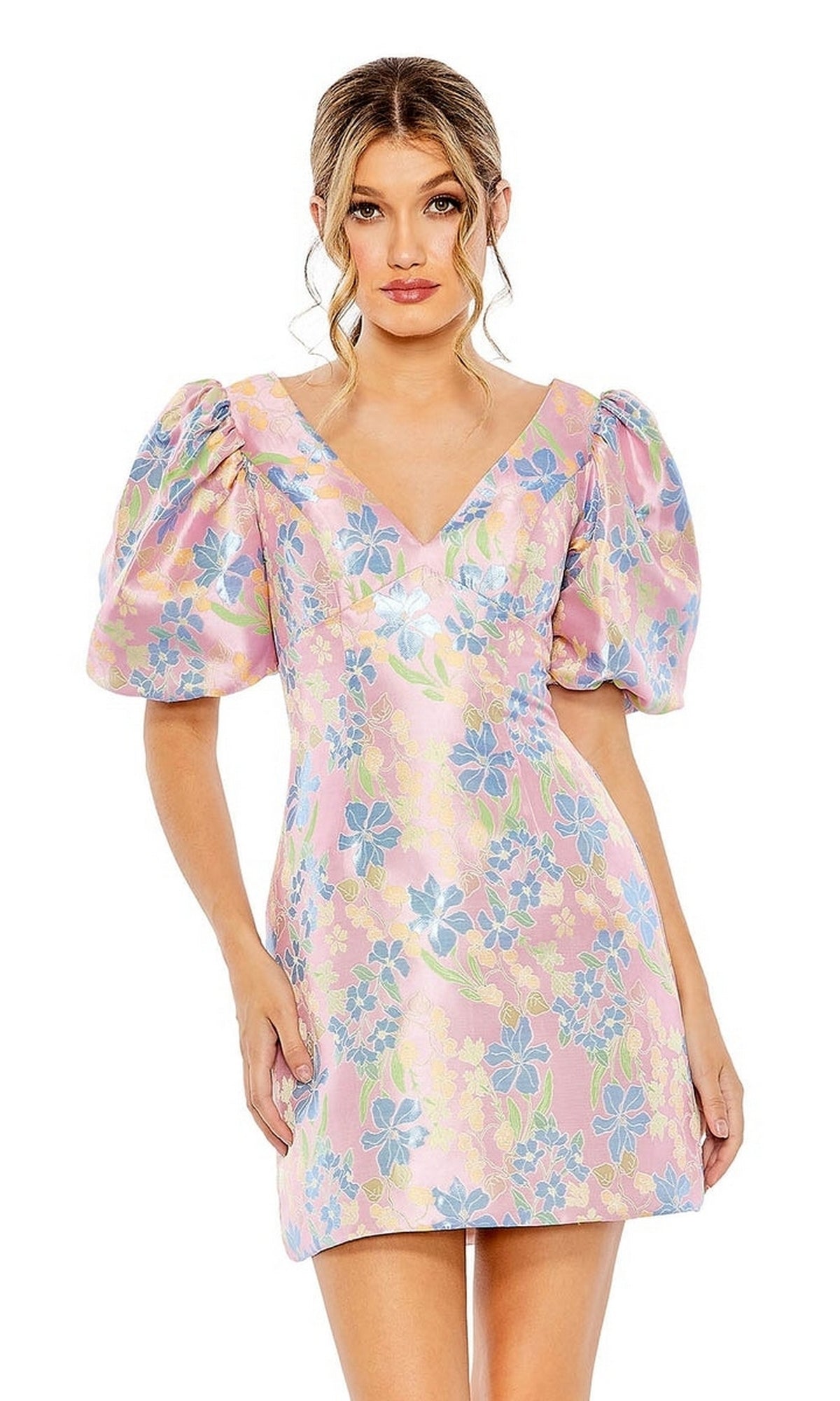A woman faces forward in the Short Homecoming Dress 49622 by Mac Duggal—a pastel pink floral print dress with blue and yellow blooms and puffed short sleeves.
