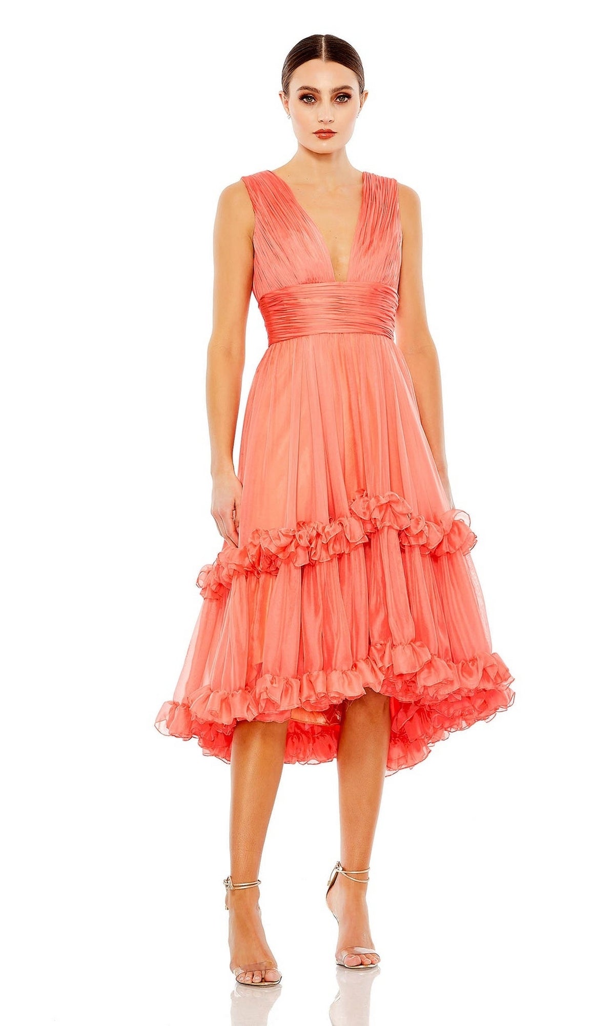A woman stands against a white background wearing the Mac Duggal Short Wedding Guest Dress 49635—a sleeveless, coral pink V-neck A-line dress with ruffled details and a high-low hem—paired with silver strappy heels.