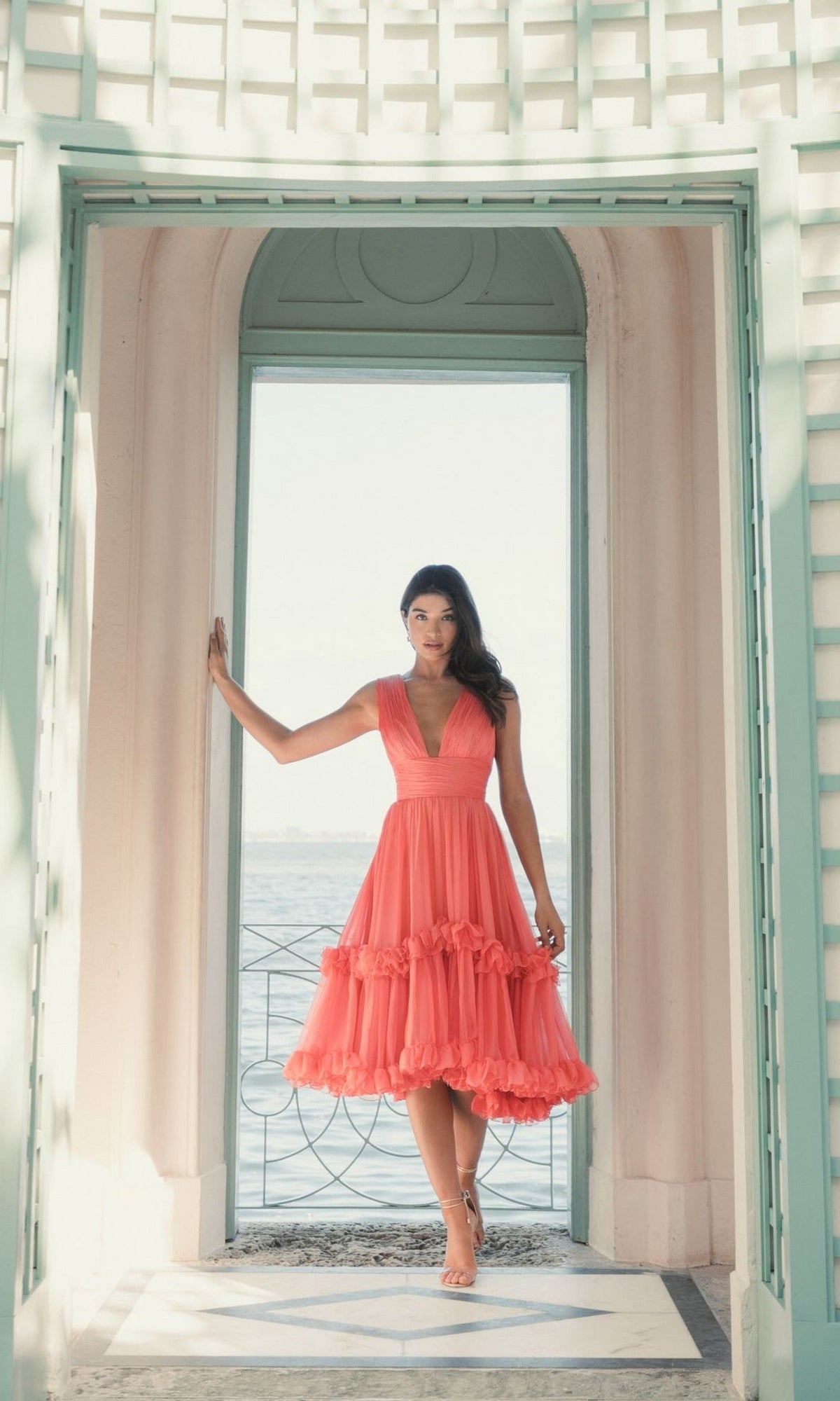 A woman in a sleeveless Short Wedding Guest Dress 49635 by Mac Duggal stands barefoot in an arched doorway, water and sky visible in the background.