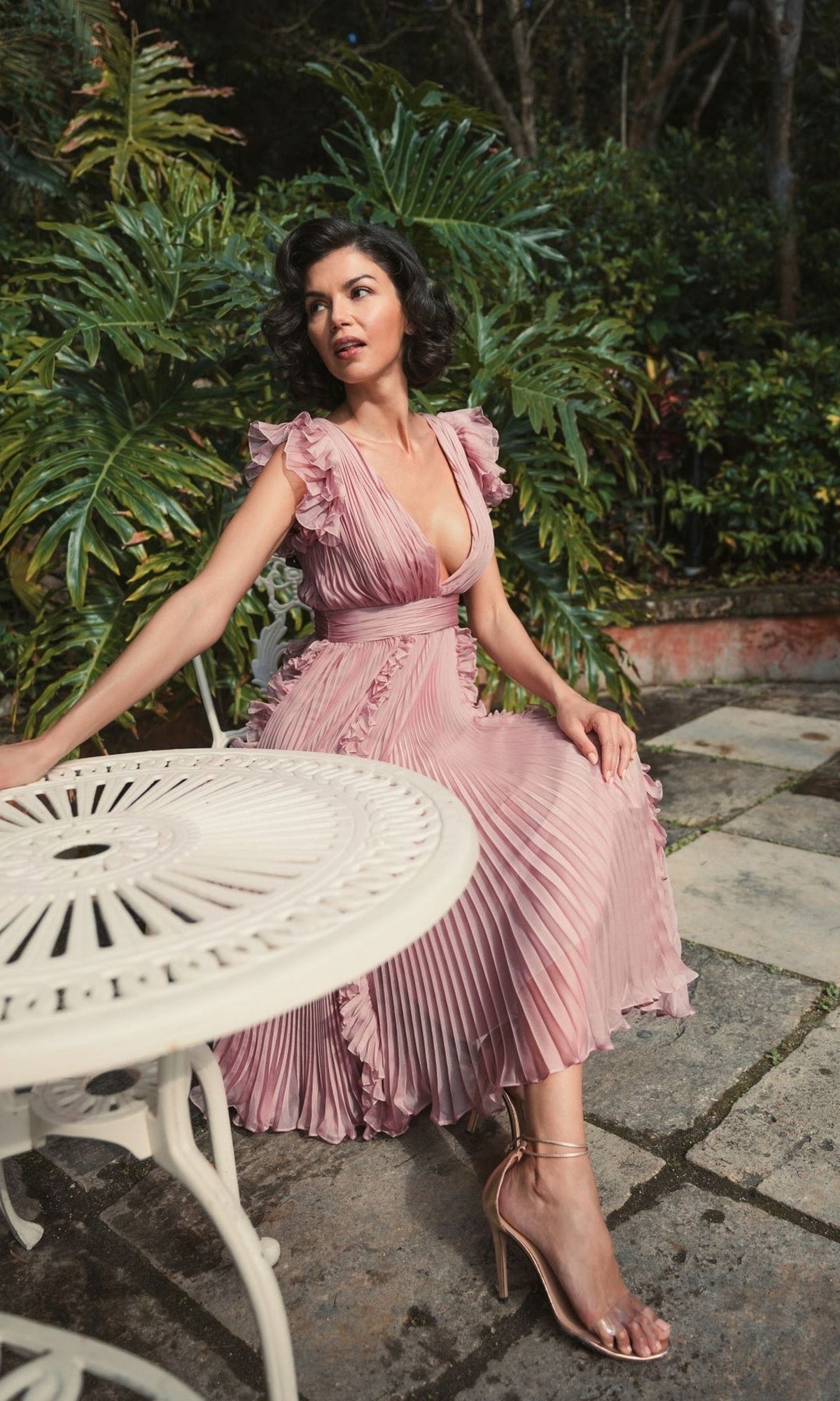 A woman in the rose pink Short Wedding Guest Dress 49675 by Mac Duggal, featuring ruffle cap sleeves and a chiffon midi design, sits on a white metal chair beside a matching table with lush green plants in the background.