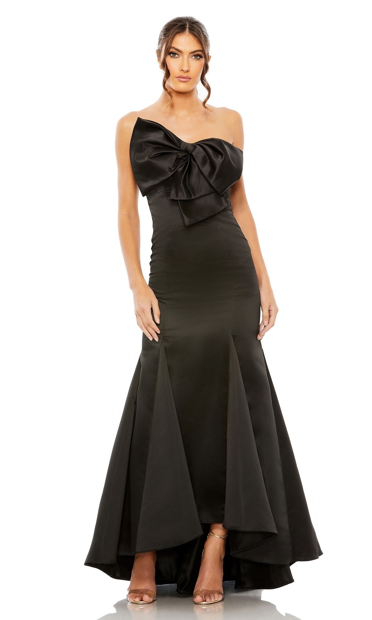 A woman wears the Mac Duggal 49701 strapless mermaid formal dress—a floor-length black gown with a large bow on the bodice, flared hem, and nude high-heeled sandals.