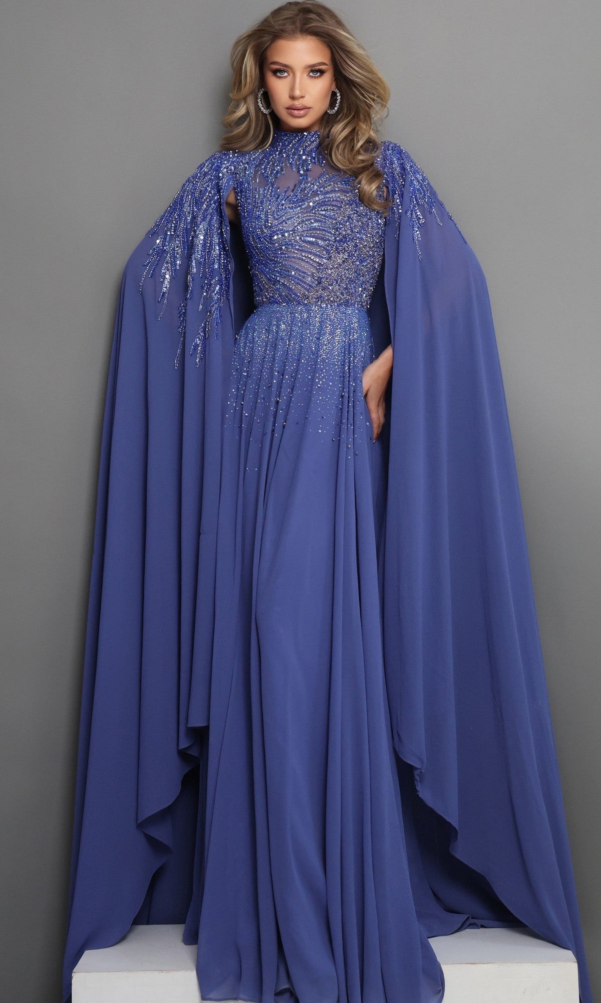 Wearing the Jovani 49742—a floor-length, royal formal dress featuring chiffon fabric, sequin embellishments, and dramatic embellished cape sleeves—a woman stands poised against a plain gray background, showcasing an elegant mother-of-the-bride look.