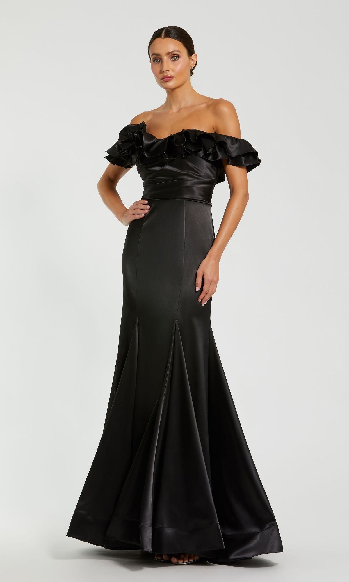 A woman wears the Mac Duggal 49834 Ruffled Long Satin Prom Dress—an off-the-shoulder, floor-length black mermaid gown with ruffled sleeves—standing against a plain light background.