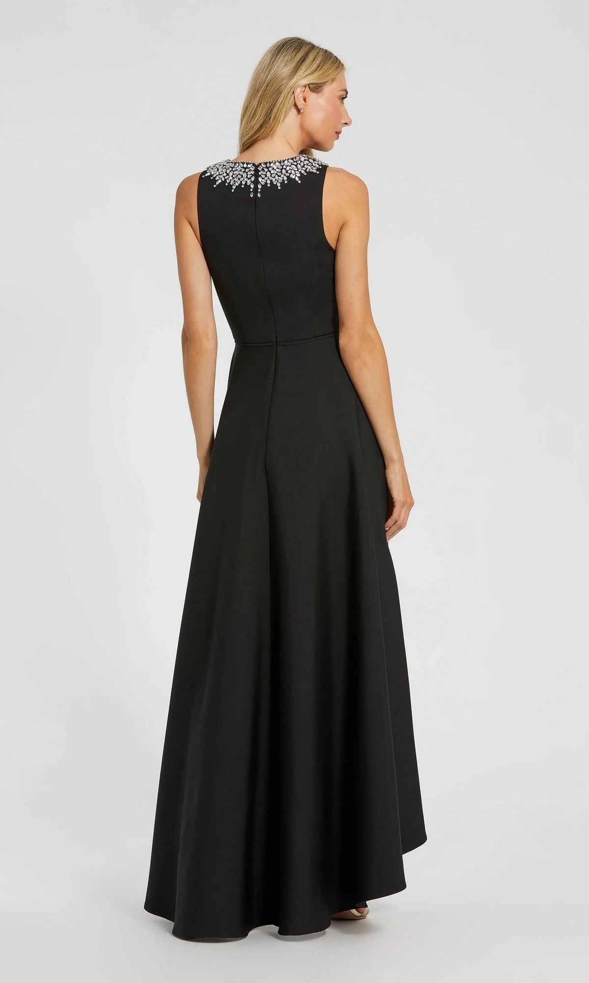 A woman stands facing away, wearing the Mac Duggal 49844 High-Low Formal Dress—a sleeveless black crepe gown with white embellishments on the upper back and an elegant a-line skirt.