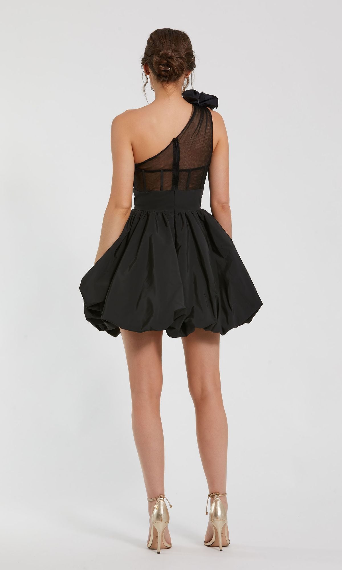 A woman faces away wearing the Mac Duggal 49858 Taffeta Short Black Party Dress, featuring a ruffled one-shoulder design, sheer panel, flared skirt, and paired with metallic high heels.