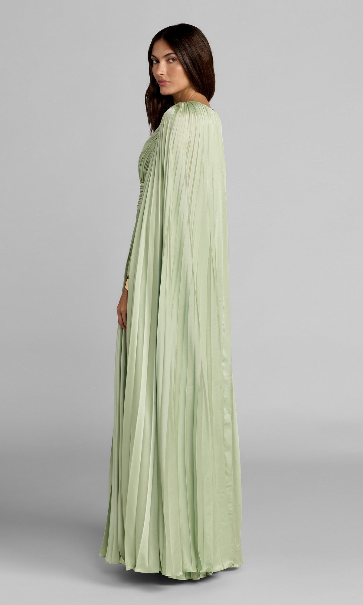 A woman stands sideways in the Mac Duggal 49872 Cape-Sleeve Long Formal Dress, featuring pleats and a cape-like back, set against a plain gray background.