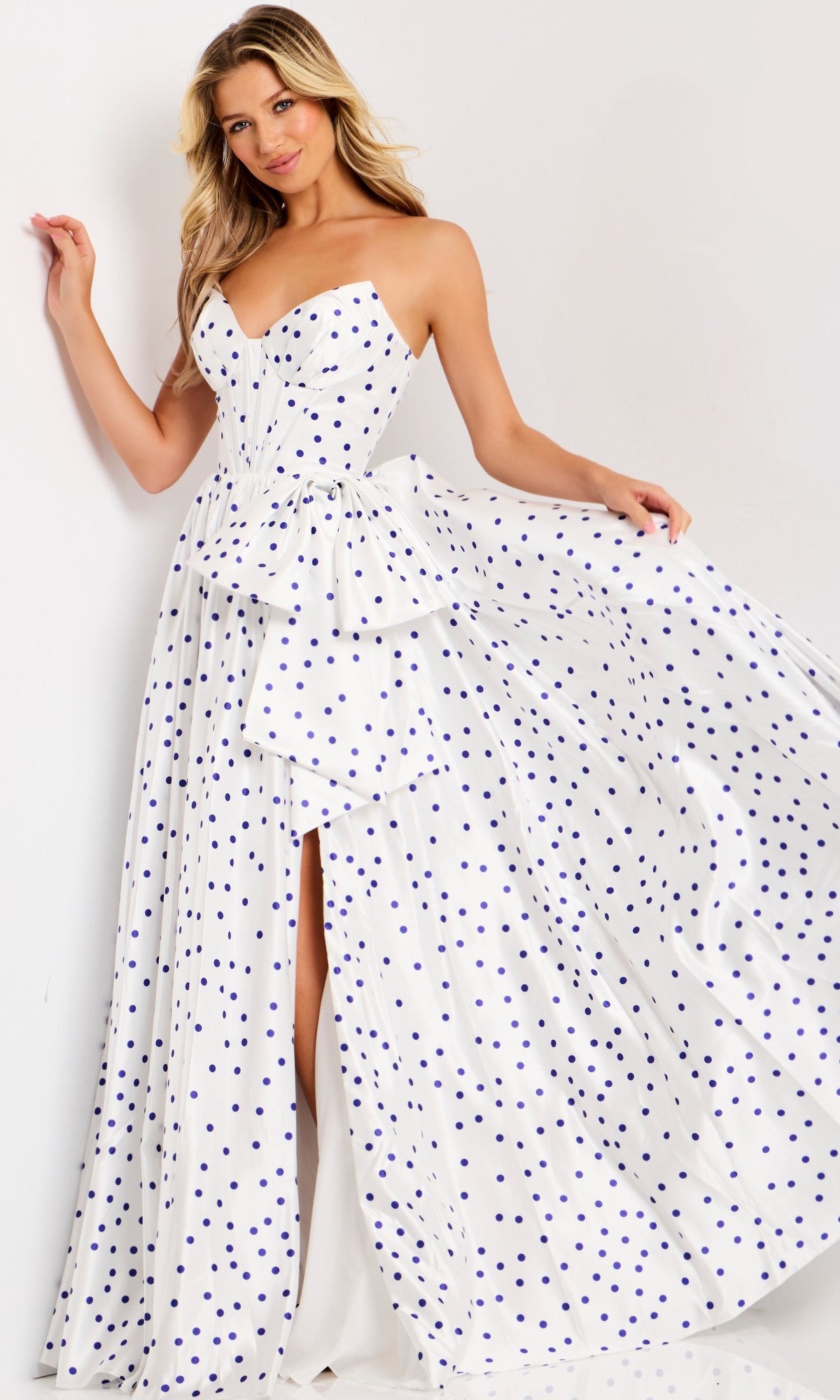 A woman stands against a plain background wearing the Jovani 49899, a long strapless polka dot prom dress with a peplum waist and thigh-high slit.