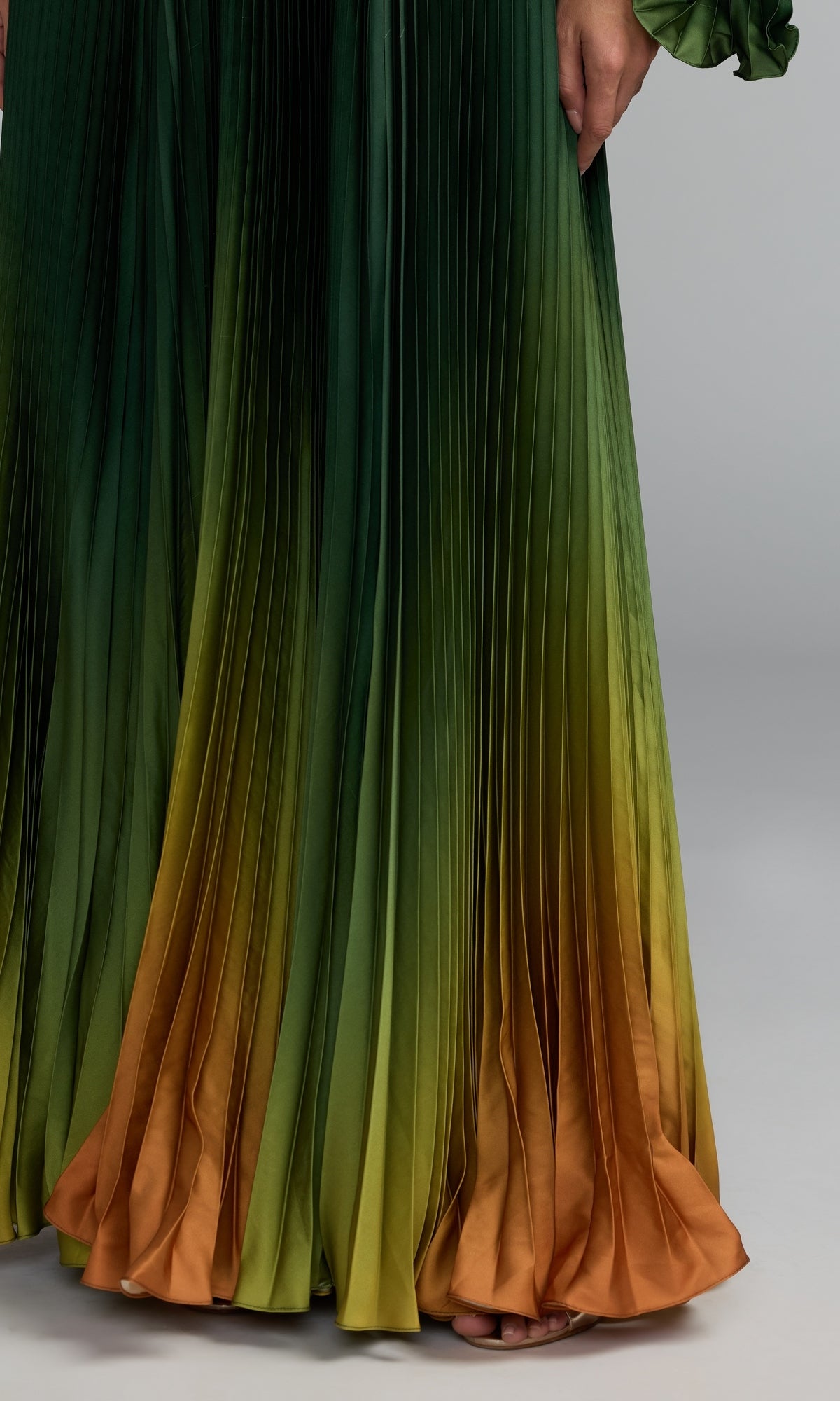 Wearing the Mac Duggal 49903 One-Shoulder Ombre Formal Dress, featuring pleated fabric in a green and orange gradient, a person stands with their hand resting at their side and bare toes peeking out at the hem.