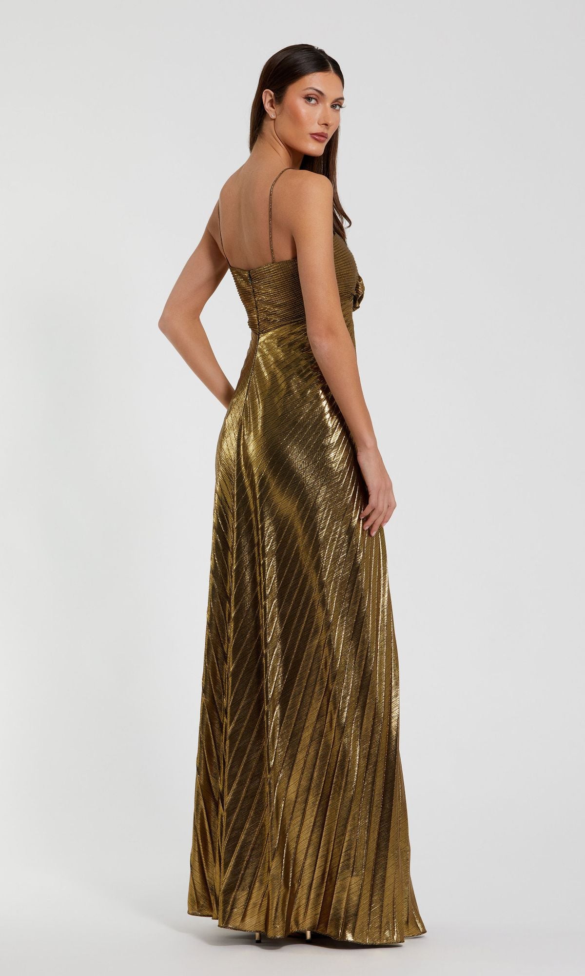 A woman models the Mac Duggal 49905 Metallic Gold Long Prom Dress, featuring metallic gold pleats and thin straps, as she poses against a plain background.