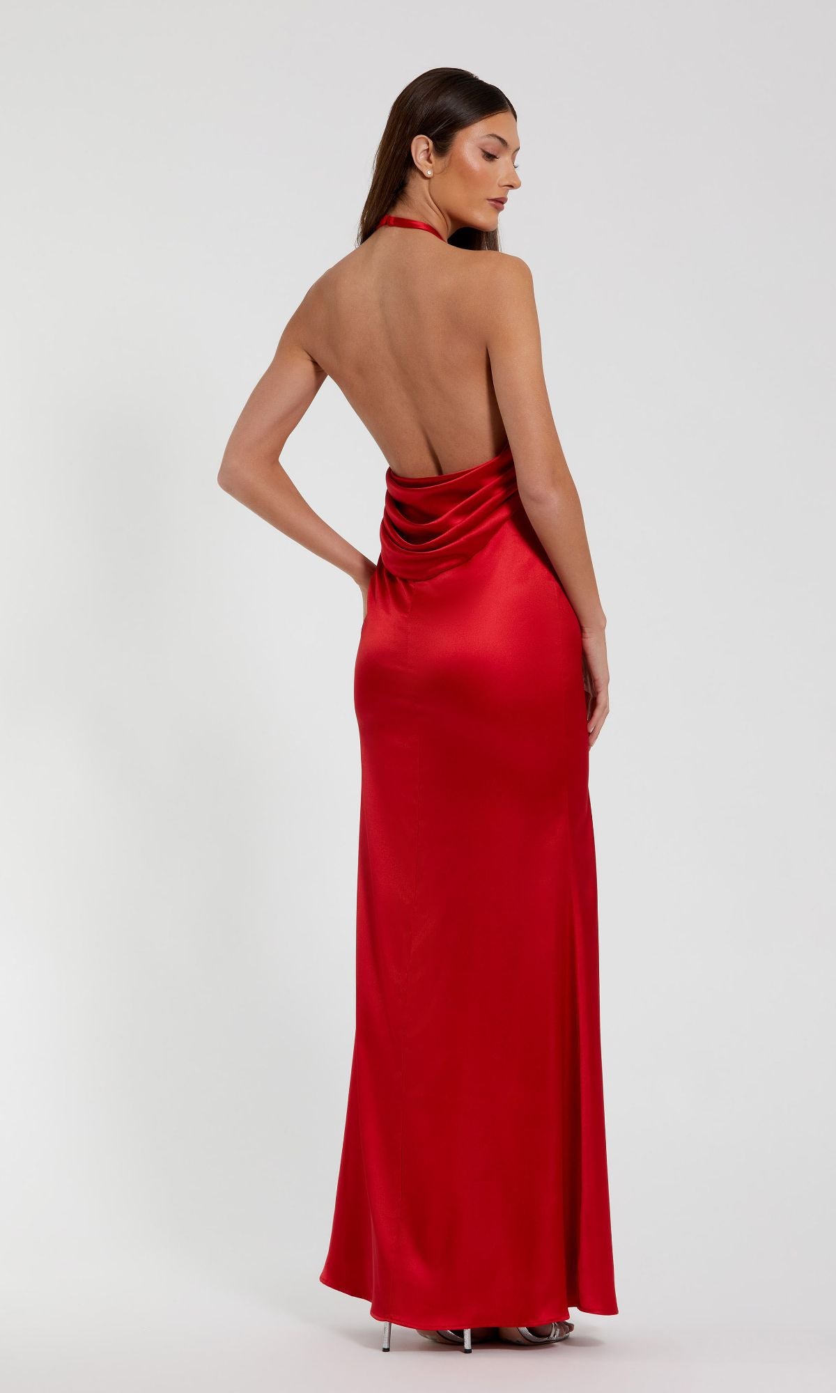 A woman in the Mac Duggal 49913 open-back long halter prom dress, featuring floor-length red satin, stands with her back to the camera against a plain background.