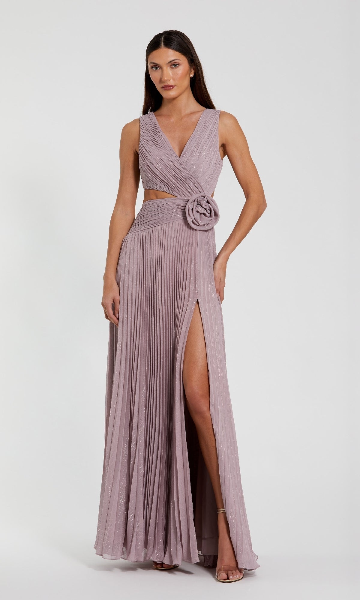 A woman stands against a plain background wearing the Mac Duggal 49938—a sleeveless mauve chiffon long formal dress featuring a v-neckline, pleated design, thigh-high slit, wrap-style bodice, and a large fabric flower at the waist.