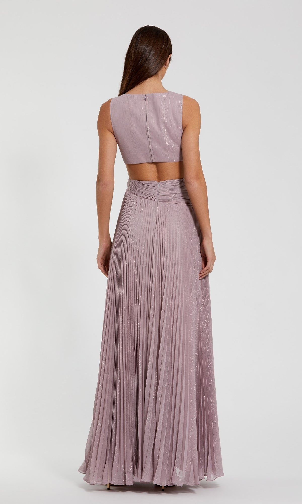 A woman with long brown hair wears the Mac Duggal 49938 Long Formal Dress, a sleeveless light mauve chiffon gown with elegant pleats and a cutout back, standing with her back to the camera against a plain background.
