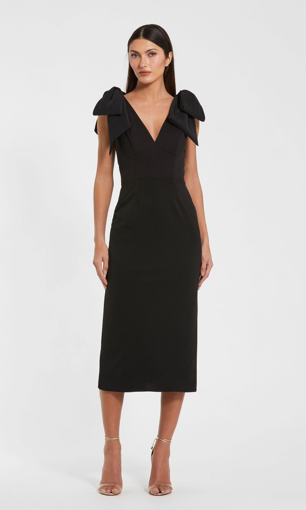 A woman stands facing forward in the Mac Duggal 49954 V-Neck Midi Wedding Guest Dress, which features a sleeveless black design with large shoulder bows, paired with strappy nude heels.