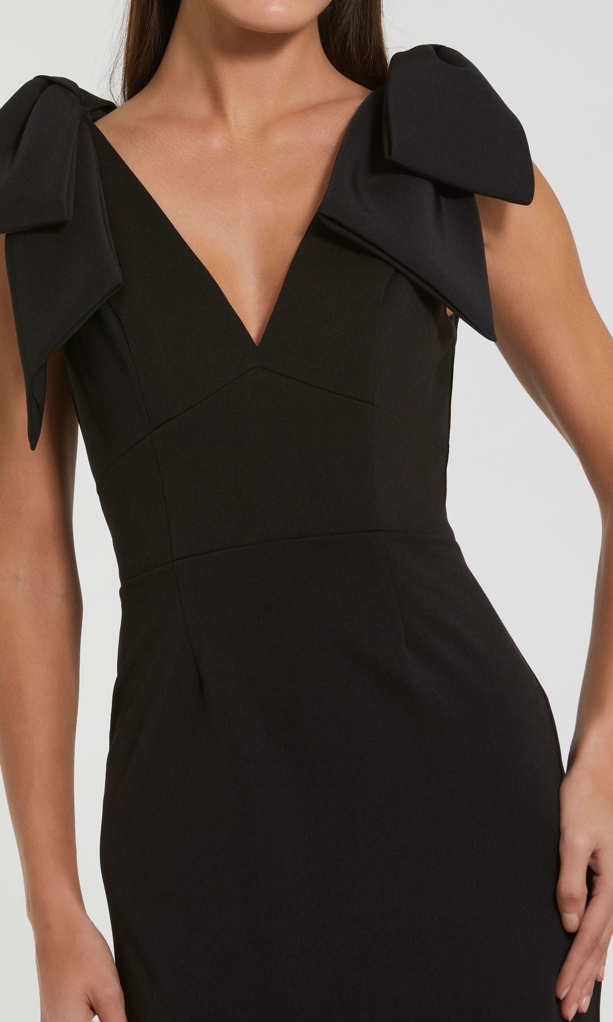 A woman models the Mac Duggal 49954 V-Neck Midi Wedding Guest Dress, a sleeveless black tea-length design featuring a deep V-neckline and large shoulder bows, posed against a plain background.