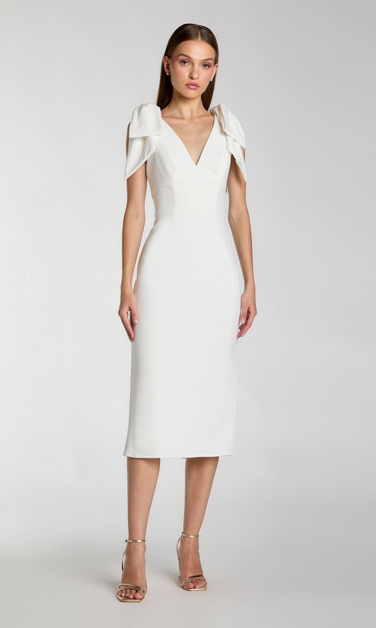A woman stands against a plain background wearing the Mac Duggal 49954 V-Neck Midi Wedding Guest Dress with draped shoulders, paired with strappy heeled sandals.