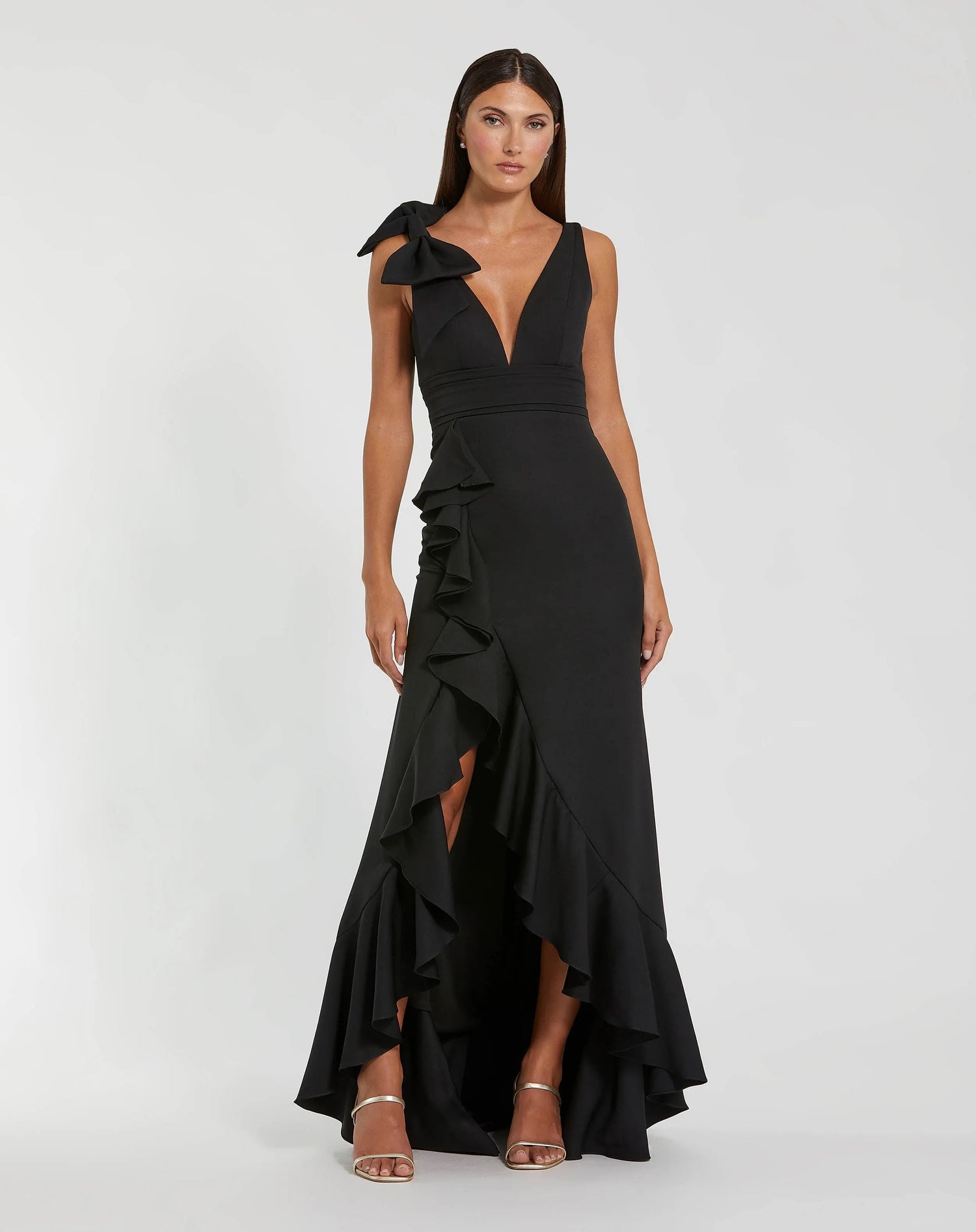 A woman stands against a plain light background, dressed in the Mac Duggal 49965 long formal dress—a sleeveless black evening gown featuring a deep V-neckline, ruffle accents, a side shoulder bow, and a striking high-low hem.