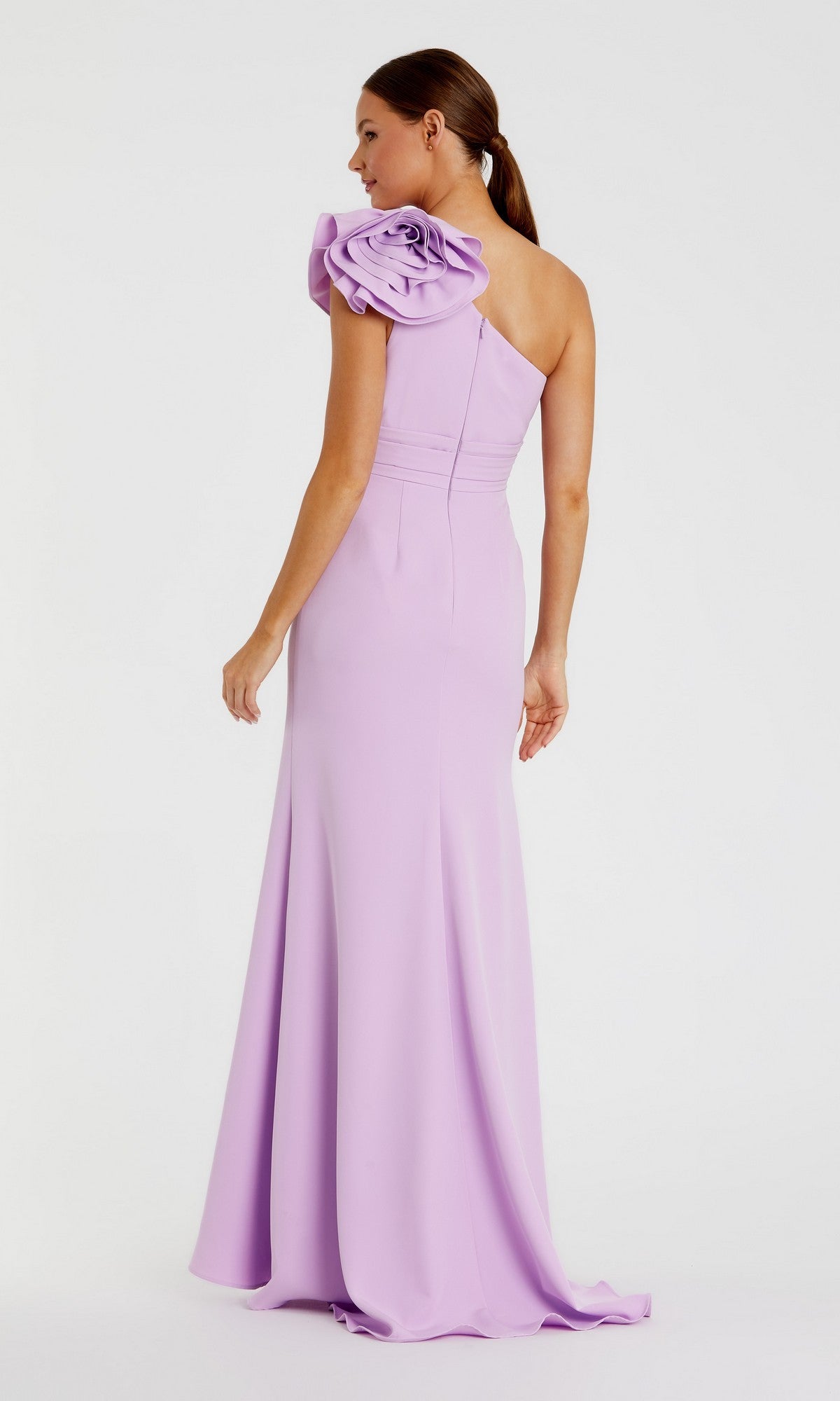 A woman stands against a plain white background, wearing the Long Formal Dress: Mac Duggal 49966—a floor-length, lilac one-shoulder formal gown featuring a dramatic fabric flower accent at the shoulder. The elegant crepe design of this dress enhances her poised silhouette and exudes sophistication.
