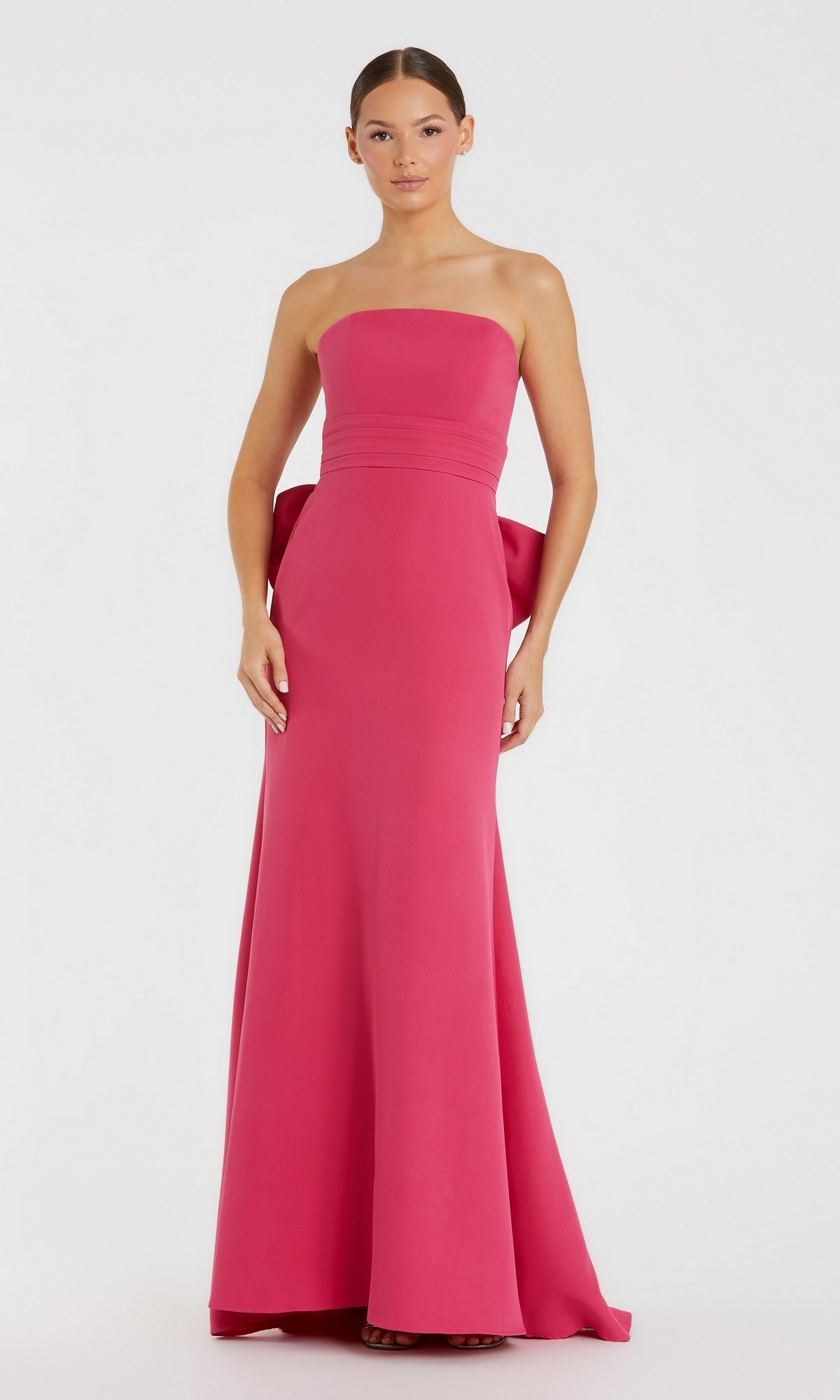 A woman stands against a plain white background wearing the Mac Duggal 49967 long formal dress in hot pink, featuring a strapless design and an oversized bow detail at the back.