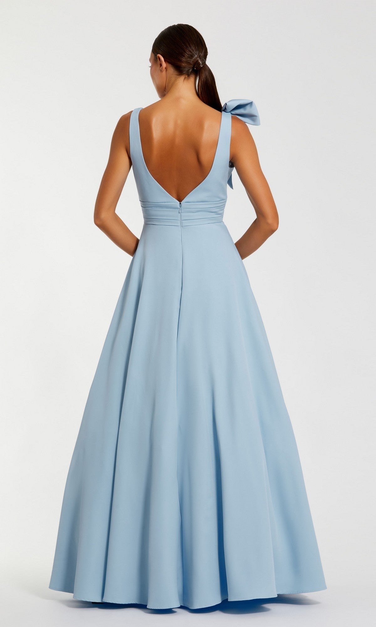 A woman faces away, wearing the Shoulder-Bow Long Formal Dress by Mac Duggal 49972—a floor-length light blue gown featuring wide straps, a deep V-back, and a large bow on one shoulder.