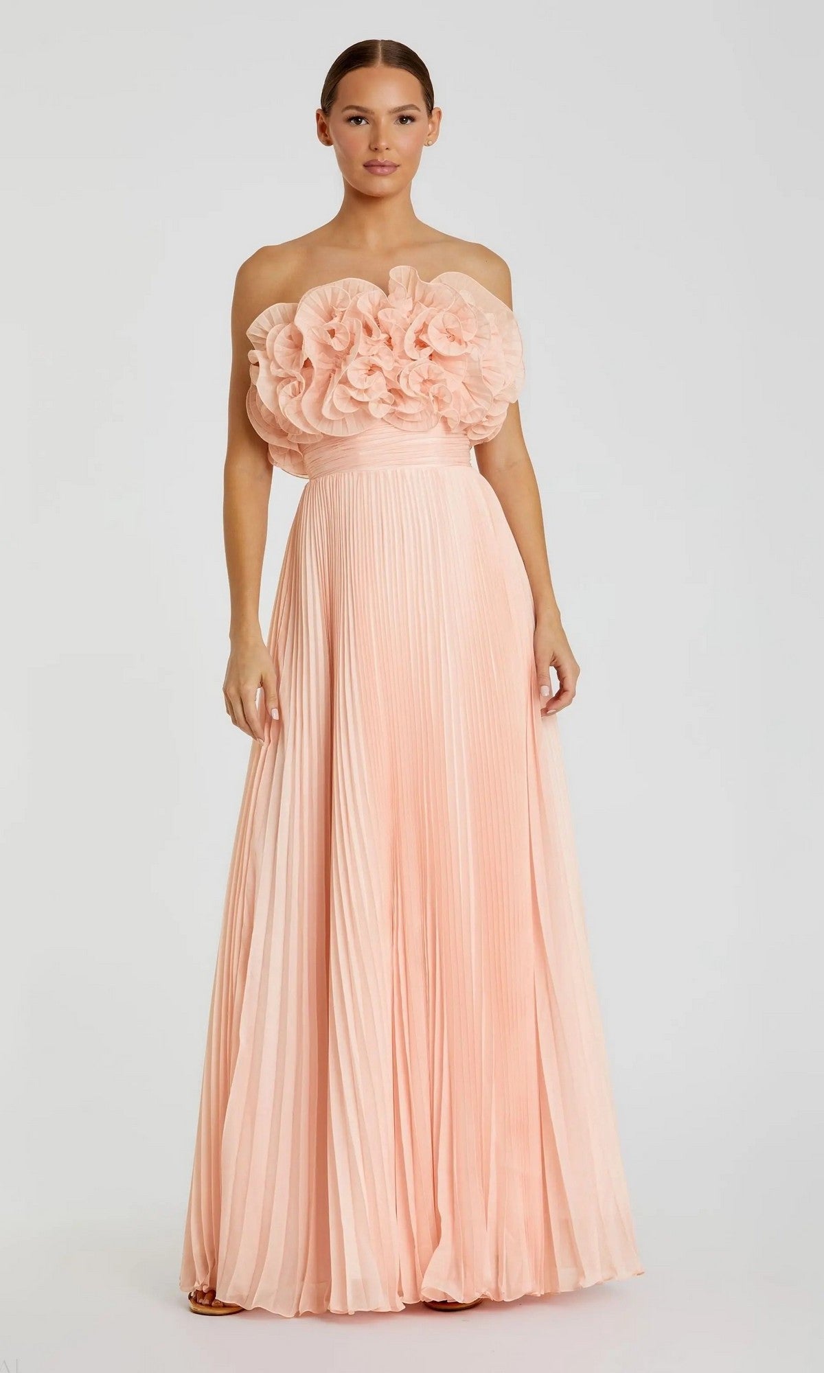 A woman wears the Mac Duggal 49975 long formal dress in blush, featuring large floral appliqués on the bodice and a pleated chiffon skirt, standing against a plain background.