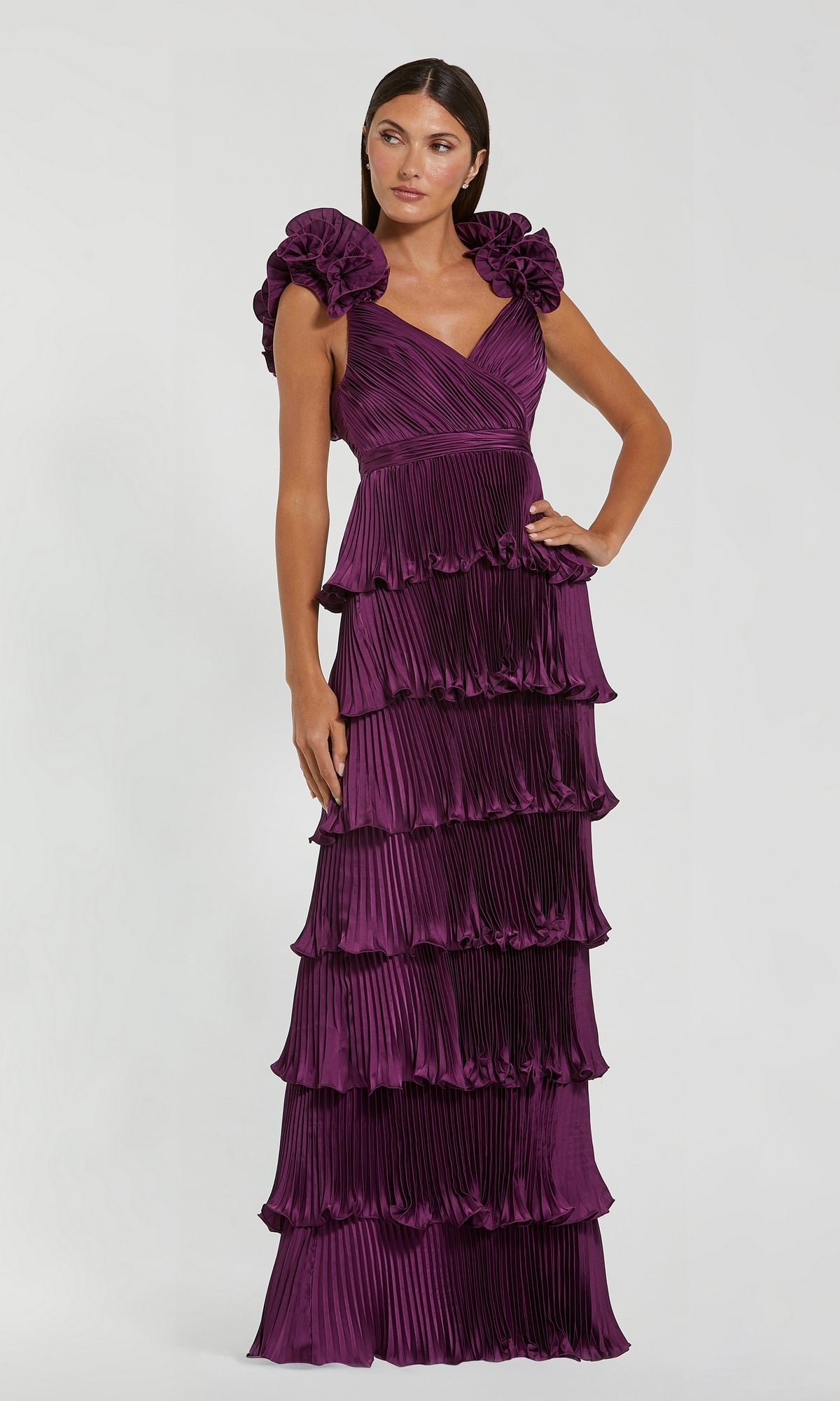 A woman wearing the Mac Duggal 49983 long formal dress in aubergine, featuring a sleeveless design with pleated fabric, ruffle shoulder straps, and a tiered floor-length skirt, stands against a plain light background.