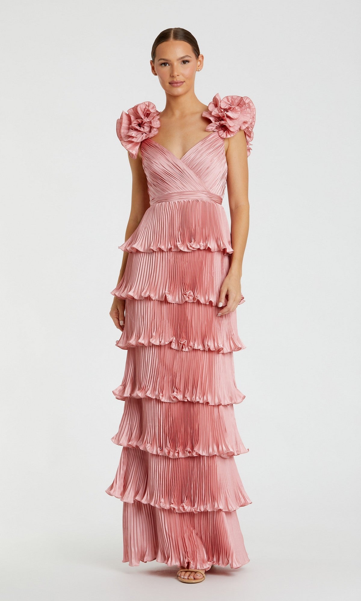 A woman stands wearing the Mac Duggal 49983 long formal dress in rose, featuring pleated fabric, a ruffle tiered skirt, and large floral shoulder details.