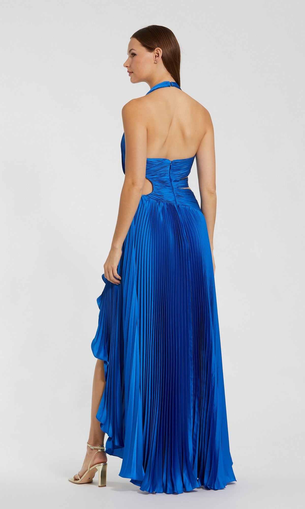 A woman stands facing away, wearing the Mac Duggal 49995 Long Formal Dress—a vibrant blue halter neck gown featuring pleats and an open back. The striking high-low hem of this dress is complemented by gold high-heeled sandals.
