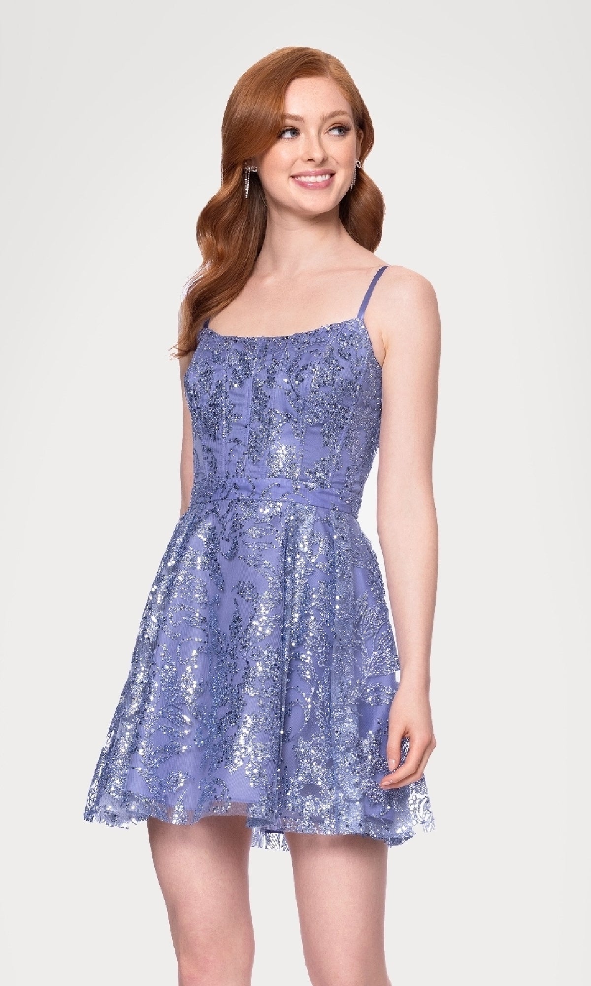 A woman stands smiling against a plain light background, wearing the Mini Glitter Hoco Dress Blondie Nites 5001BN-1— a sleeveless, lavender cocktail dress with spaghetti straps and subtle glitter.