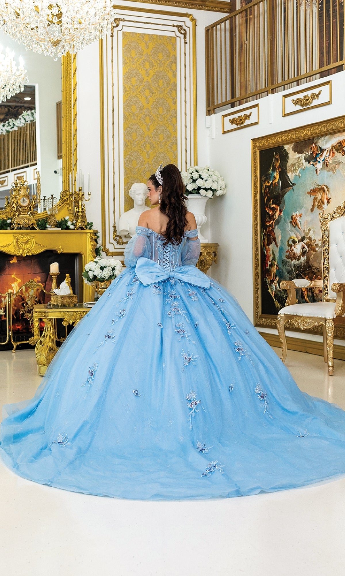 A woman wearing the Quinceanera Dress 5005 By Dancing Queen, featuring a blue off-the-shoulder sweetheart neckline, stands facing away in an ornate room with gold accents, a fireplace, flowers, and classical art.