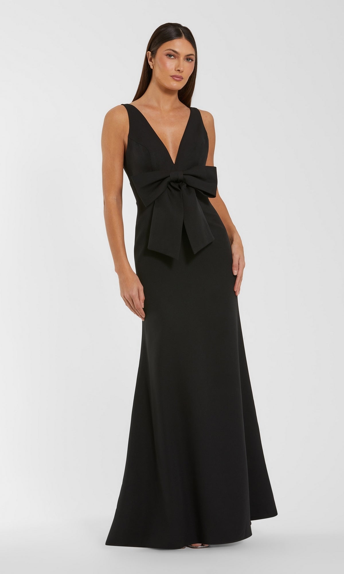 A woman stands against a plain white background wearing the Bow-Front Formal Dress: Mac Duggal 50087 in black, a floor-length evening gown featuring a large front bow, sleeveless design, and deep V-neckline. This elegant long formal dress radiates timeless sophistication.
