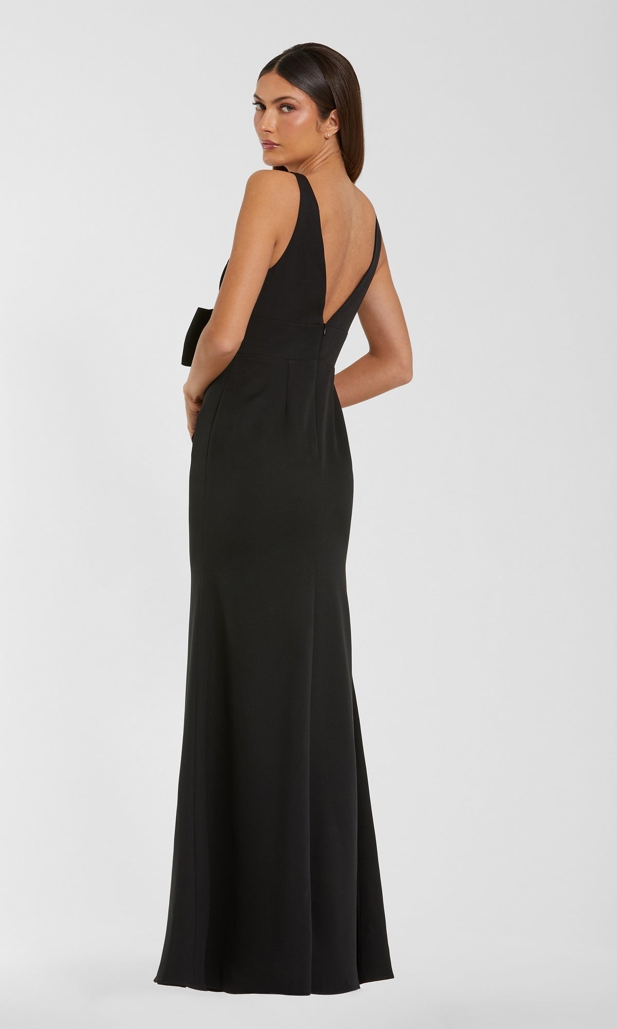 A woman stands with her back to the viewer, glancing over her shoulder in a sleeveless, floor-length black Bow-Front Formal Dress: Mac Duggal 50087, featuring a visible zipper on the back.