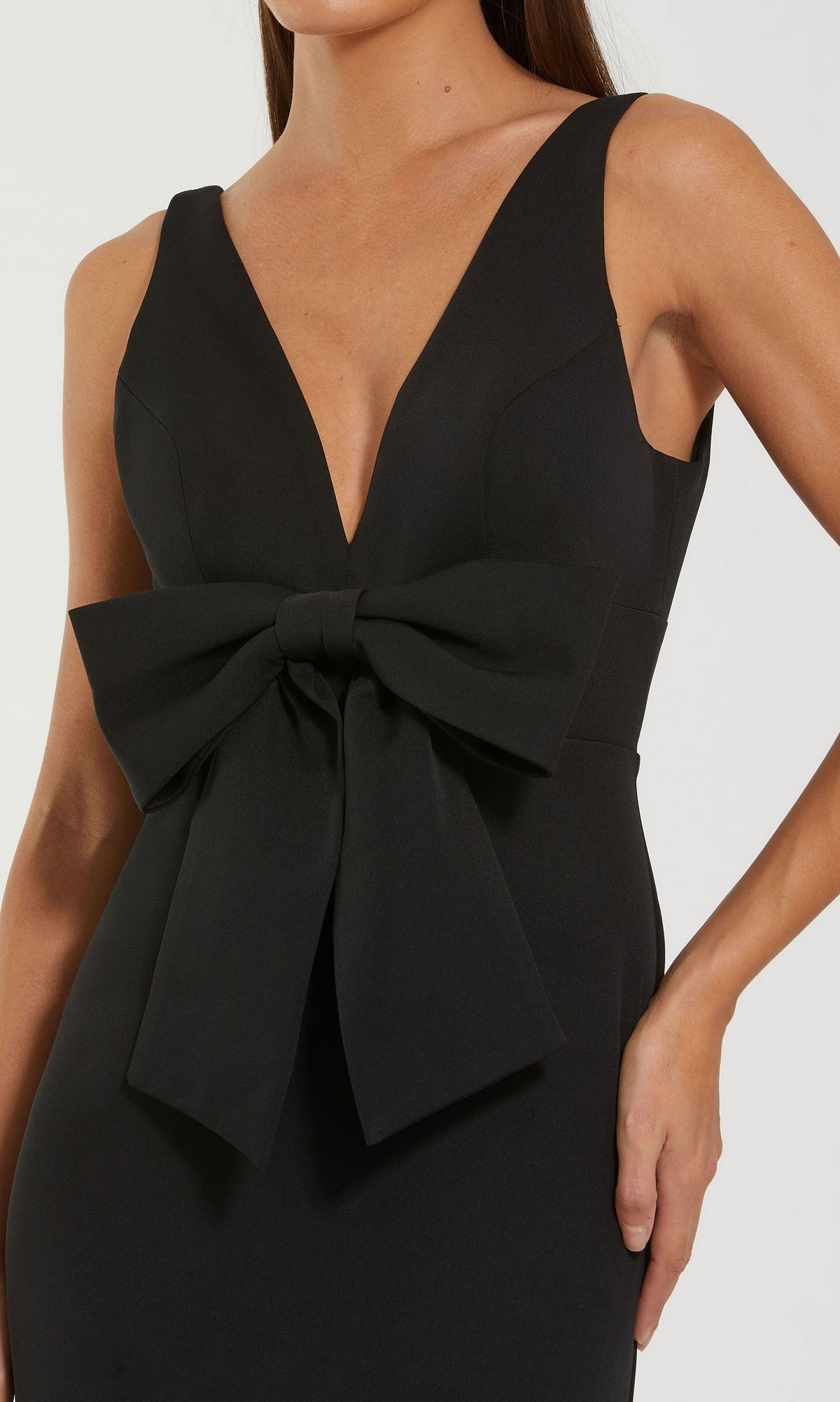 Woman wearing the Bow-Front Formal Dress: Mac Duggal 50087, a sleeveless black gown featuring a deep V-neckline and a statement bow detail at the waist, posing with one hand on her hip.